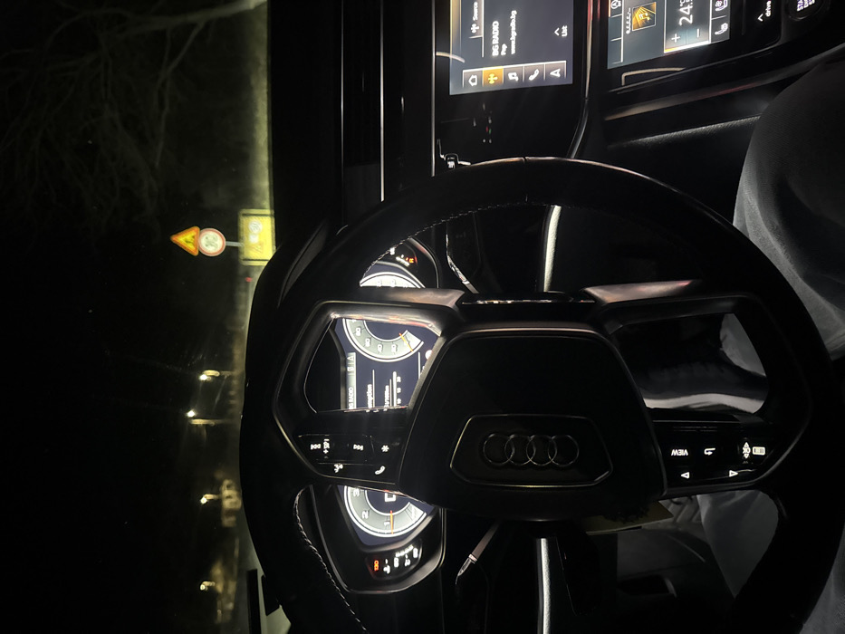 The image depicts the interior of a vehicle, specifically the driver's perspective looking out at night in the city of Banichan, Bulgaria. In the foreground, a black, multi-functional steering wheel with a prominent Audi logo is centered. Behind the steering wheel, the digital instrument cluster is illuminated, displaying various gauges and information, including a "0.0 l/100km" consumption reading and a digital clock. To the right of the steering wheel, a portion of the digital infotainment screen is visible, showing "BG RADIO," "Pop," and "www.bgradio.bg," along with a climate control display set to "24°C." The driver's lower torso and legs, clad in light-colored pants, are partially visible below the steering wheel.
Through the windshield, the dark exterior environment of Banichan, Bulgaria, is illuminated by distant streetlights. Visible objects include a yellow triangular road warning sign superimposed over a circular white sign with a red border, indicating a "30" speed limit or similar restriction, positioned above a rectangular yellow sign. Bare tree branches are discernible against the dark sky. The scene suggests the vehicle is stationary or moving very slowly at night.