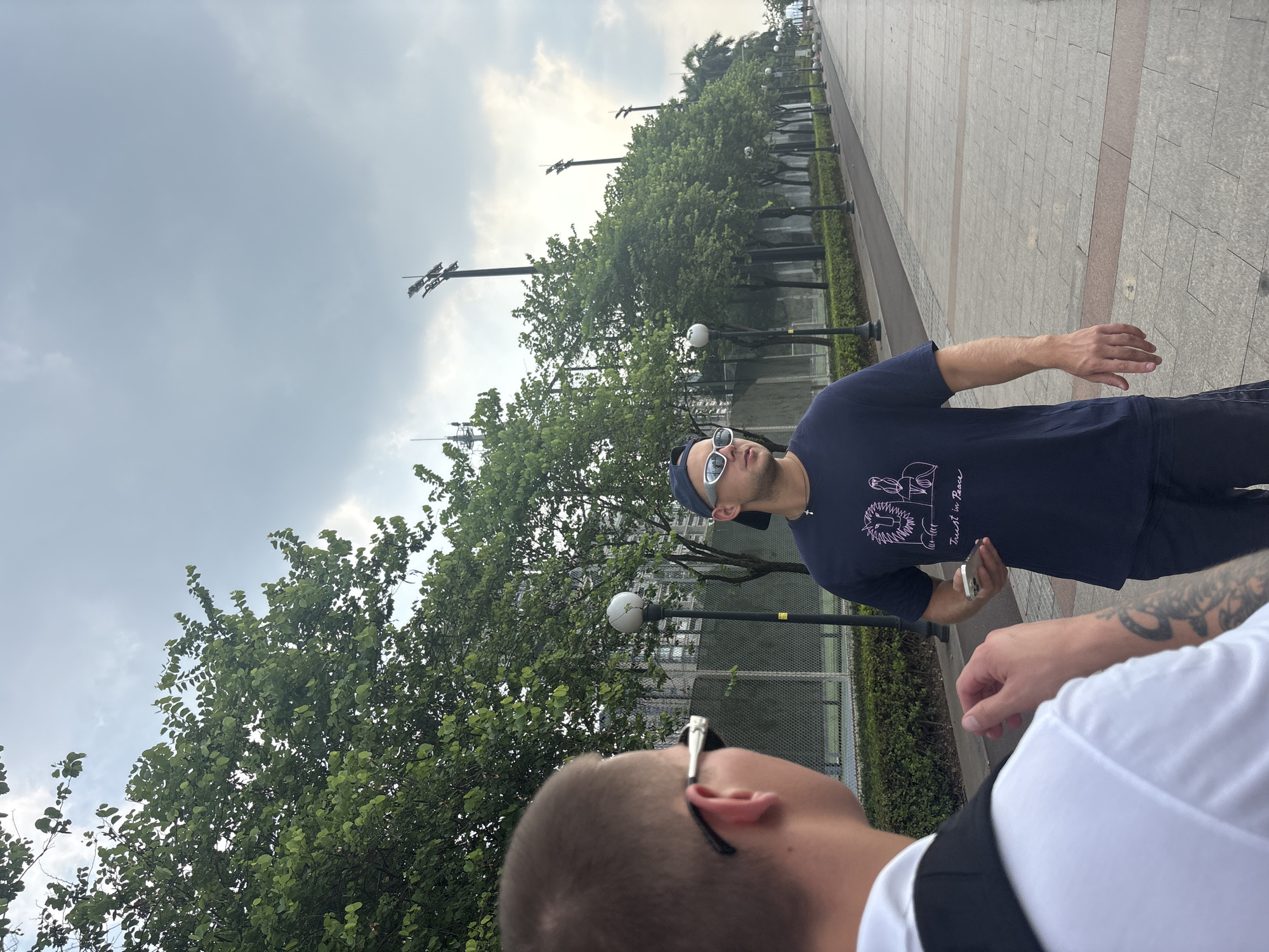 Two men are standing on a paved walkway in Haizhu District, China, with a row of trees and streetlights lining the path to the right. The man in the foreground, seen from behind, is wearing a white shirt and has tattoos on his arm. The man in the background is facing left, wearing a dark blue t-shirt with a graphic design and text that reads "Trust in Peace," a blue baseball cap, and sunglasses. He is holding a smartphone in his right hand. The walkway is paved with large rectangular tiles and has a curb on the left separating it from a grassy area with bushes and a chain-link fence. In the distance, there are tall light poles with multiple lamps, and buildings can be seen behind the trees. The sky is overcast with scattered clouds.