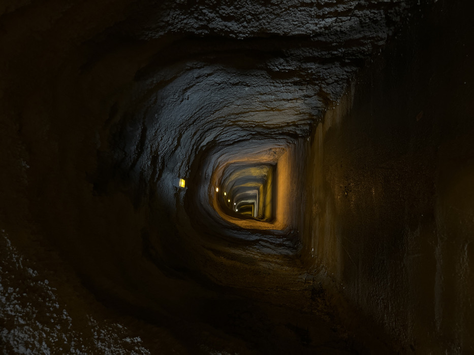 The image displays the interior of a man-made tunnel in Rafailovići, Montenegro, extending into the distance with a vanishing point perspective. The tunnel walls and arched ceiling are constructed from rough, uneven rock or concrete, exhibiting varied textures and shades of dark grey and brown. A series of evenly spaced, warm-toned, rectangular light fixtures are mounted along the ceiling, emitting an orange-yellow illumination that brightens the tunnel's interior and highlights the textured surfaces. Approximately seven or more such lights are visible, progressively illuminating the passage further down. The tunnel floor is a dark, uneven path, appearing damp or reflective in areas directly beneath the light sources. No individuals, vehicles, or other distinct objects are present in the scene.