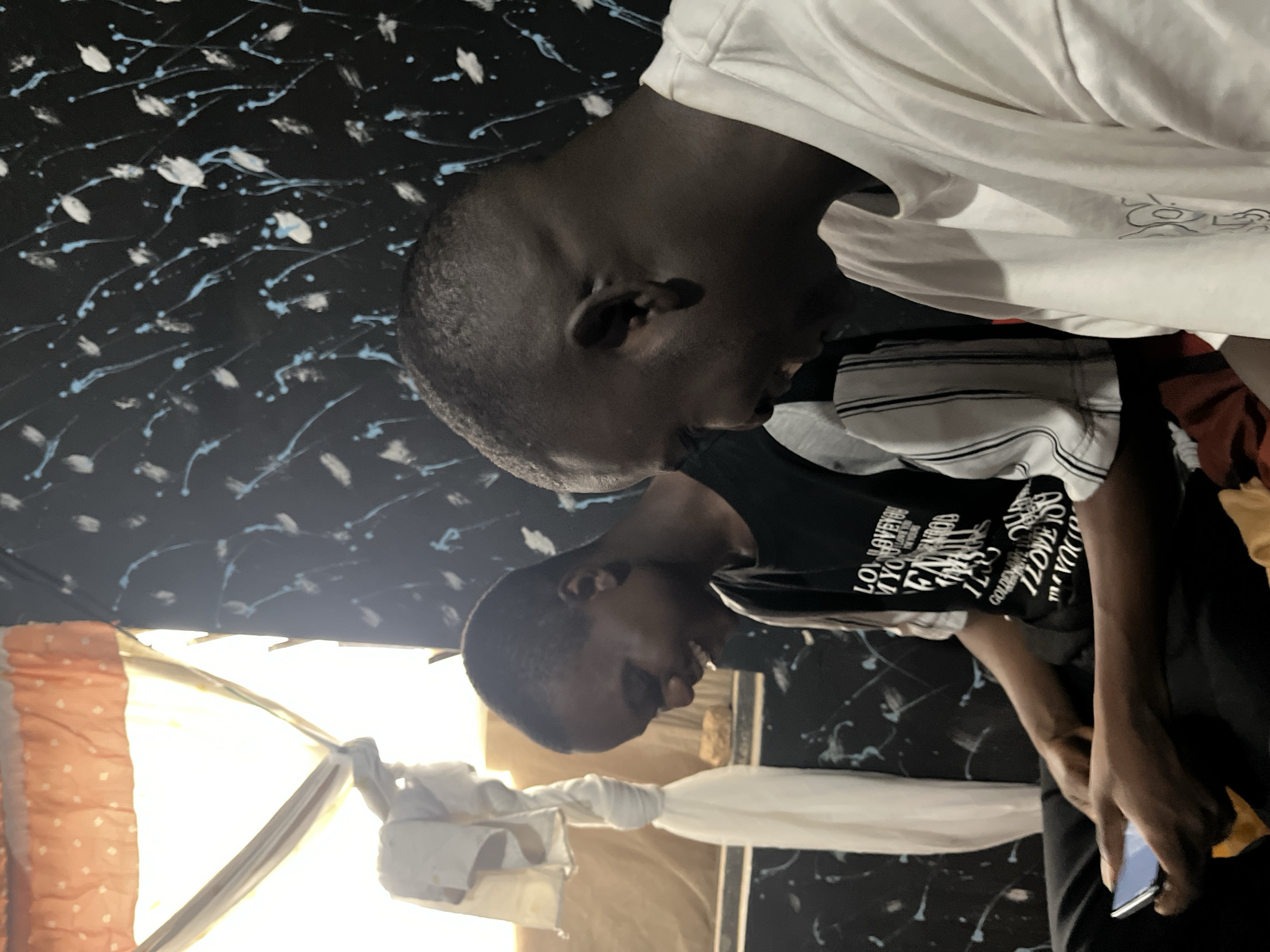 The image depicts an indoor scene with two young men seated. The man on the left, facing left, is wearing a black t-shirt with white lettering and is holding a smartphone in his left hand. He appears to be laughing. The man on the right, facing left, is wearing a white t-shirt and also appears to be smiling. The background features a black wall with white and blue abstract paint splatters, and a window covered by a patterned orange fabric and white cloth drapery. The overall setting suggests a casual indoor environment, possibly a residential room in Jos, Nigeria.