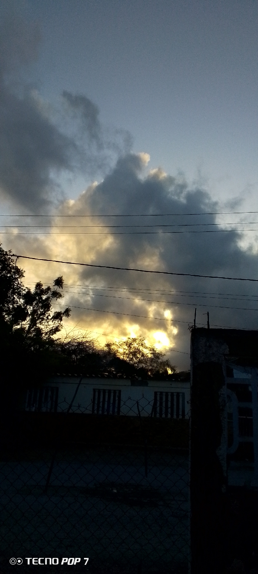 A low-angle shot captures a dusky sky with dramatic clouds illuminated by the setting sun. Power lines crisscross the sky, silhouetting the scene below. In the middle ground, a dark mass of trees obscures a low-rise building with barred windows. A chain-link fence runs across the foreground, partially obscuring the view. The bottom left corner of the image features a "TECNO POP 7" watermark.