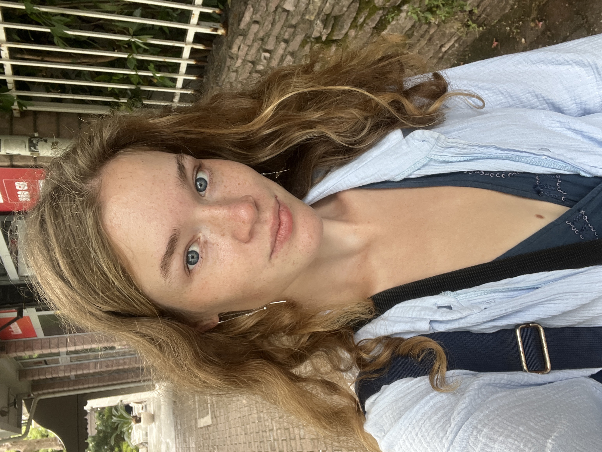 A light-skinned female with light brown wavy hair and blue eyes faces the camera. She has noticeable freckles on her face. She wears a light blue shirt over a darker blue top. A dark blue strap crosses her body. The background includes a white metal fence, a stone wall, and a glimpse of buildings and greenery, indicating an urban environment. The photo appears to have been taken in Budva, Montenegro.