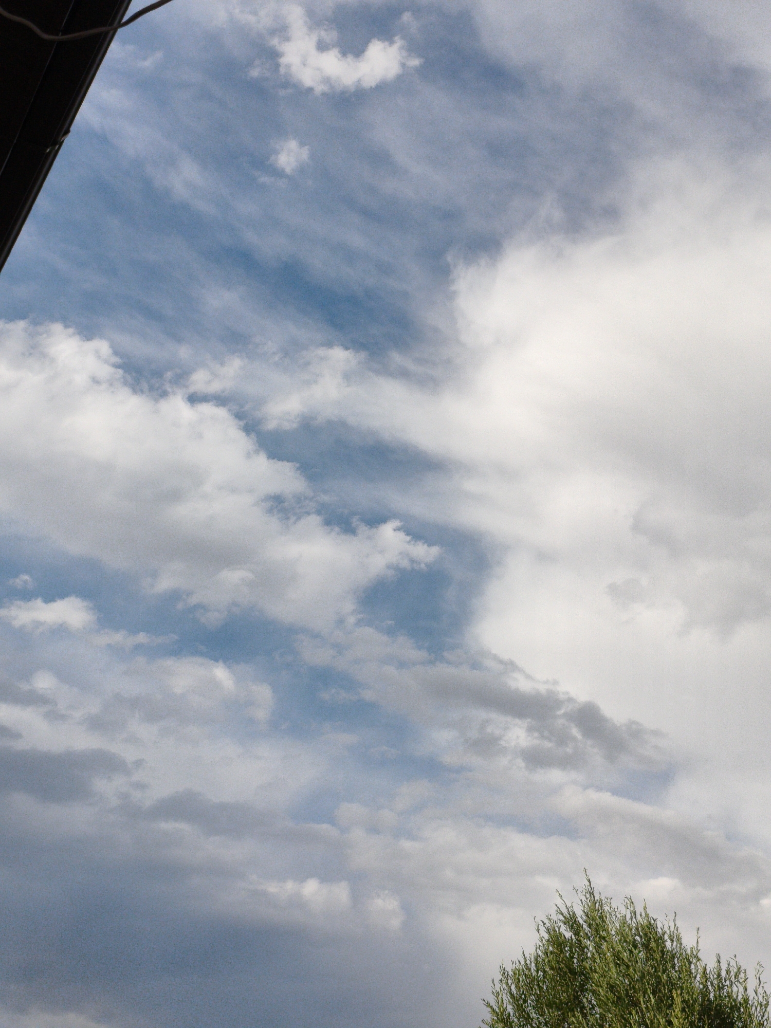 The image shows a sky with clouds and a tree. The sky is blue with white and gray clouds. Some clouds are wispy, while others are puffy. The tree is green and leafy and is in the bottom right corner of the image. A dark structure is visible in the top left corner, possibly a roof or an overhang.