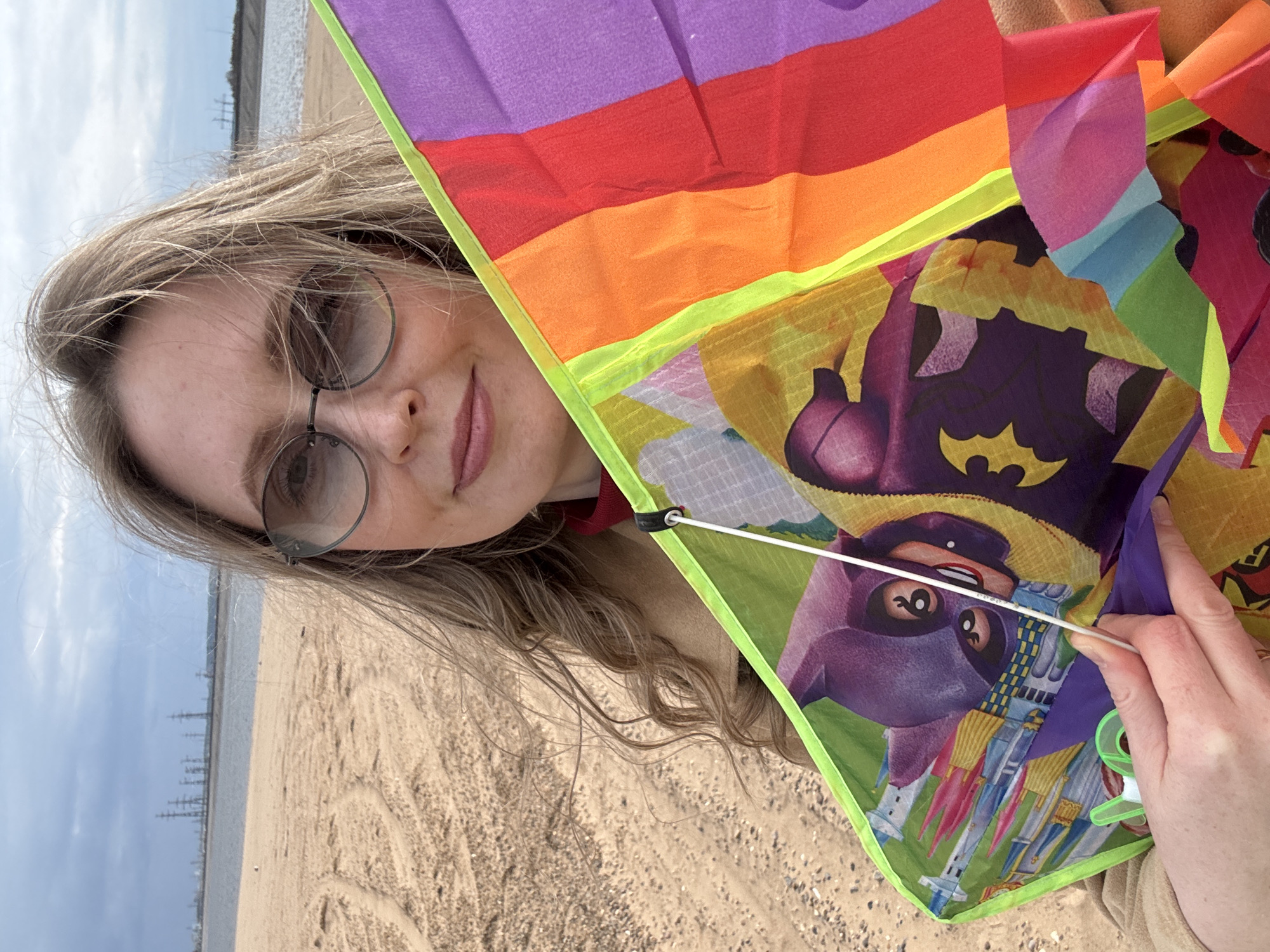 A young adult Caucasian woman with light brown hair and round glasses is shown at an angle, holding up a bright-colored kite with a Batman design. The kite also features rainbow stripes. The woman is smiling slightly. The background shows a sandy beach, water, and industrial structures in the distance, suggesting a location in or near Noviy, Russia.