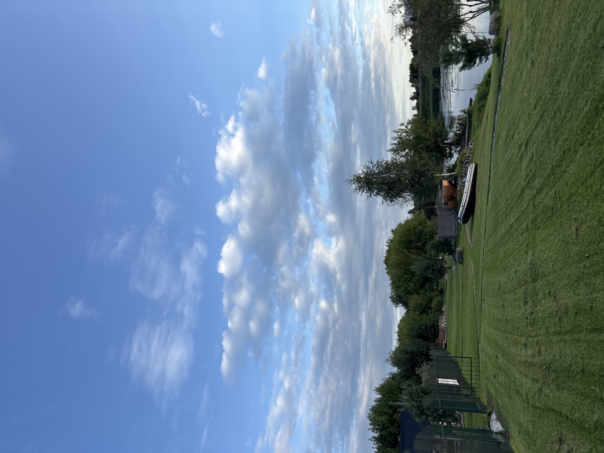 The image depicts a serene natural landscape on the outskirts of Zvenigorod, Russia, with a focus on a meticulously maintained grassy area leading down to a tranquil body of water. The sky is a vibrant blue, adorned with scattered white and gray clouds, suggesting a pleasant day. A dark green metal fence runs along the left side of the foreground, bordering a neatly mowed lawn. Several trees and bushes populate the middle ground, with a distinctive tall birch tree standing out. Near the water's edge, an inflatable boat rests on the grass, alongside a small wooden structure, possibly a sauna or shed, and a portion of what appears to be a wooden deck or dock. The water reflects the sky, creating a calm and inviting atmosphere. In the distance, the opposite bank is lined with trees, and faint outlines of buildings are visible on the horizon, indicating the proximity of a settlement. The overall impression is one of peaceful, rural life by the water.
