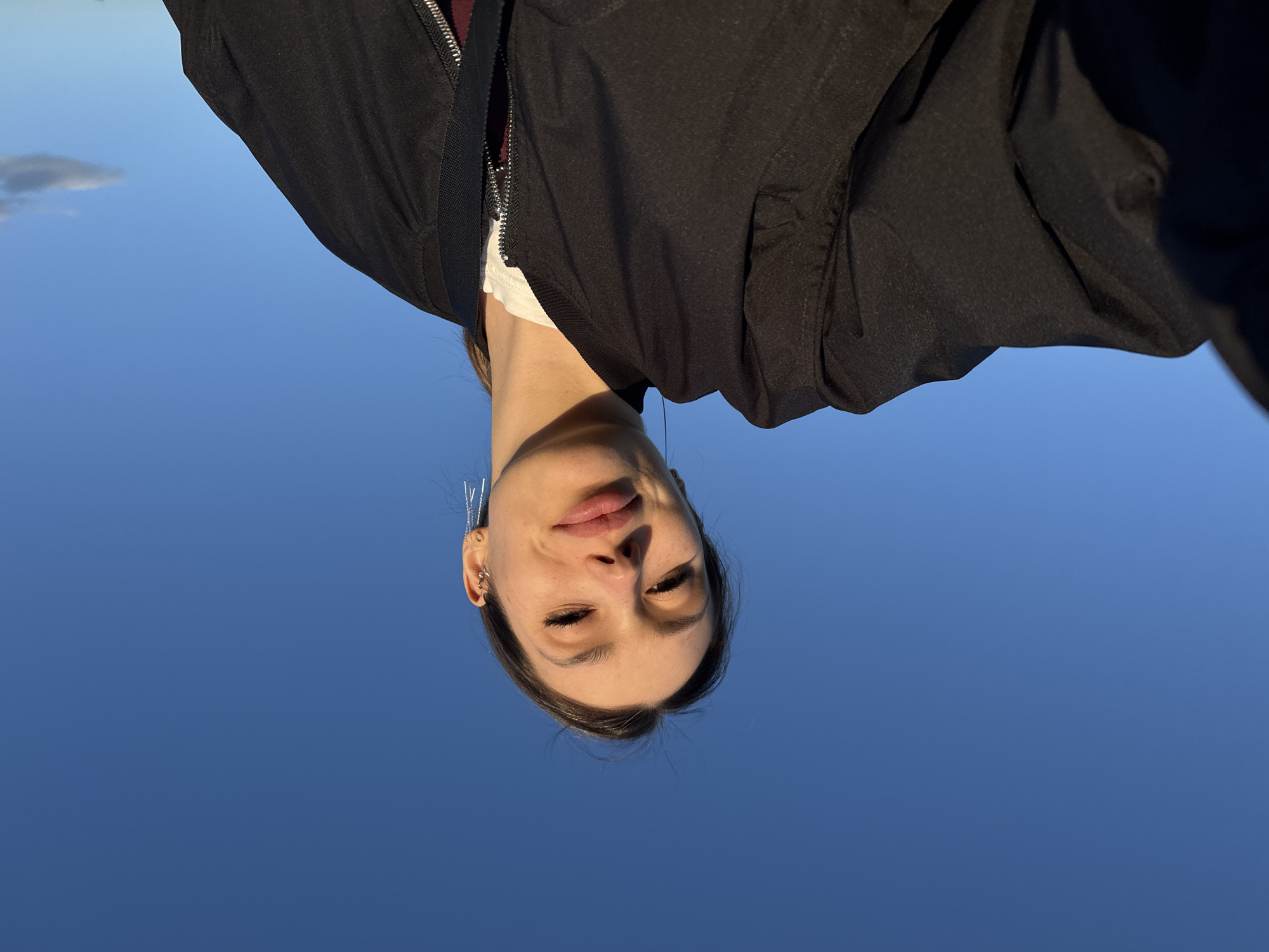 An upside-down portrait features a person, likely female, captured from a low angle against a clear, bright blue sky. The individual's head is positioned at the bottom of the frame, facing upwards, with their body extending towards the top.
The subject has fair to medium-toned skin, dark hair, and closed or partially closed eyes, giving a serene or contemplative expression. Their lips are slightly parted, revealing natural color. Multiple ear piercings, including small hoops and stud-like adornments, are visible on the right ear (from the viewer's perspective). Some silver, chain-like accessories or hair strands are visible near the top of their head, extending into the sky.
They are wearing a black bomber-style jacket with a prominent silver zipper, which is partially open, revealing a white garment underneath, and a hint of a dark red or burgundy layer beneath the white one. The fabric of the jacket appears slightly textured and matte.
The background is almost entirely a vibrant, cloudless blue sky, suggesting an outdoor setting during the daytime. A small, wispy white cloud is visible in the bottom-left corner of the image (which would be the upper-left if the image were rotated correctly). The lighting is bright and natural, casting subtle shadows under the jawline and neck, indicating sunlight from above. No text is visible in the image. The unusual orientation suggests it might be an inverted selfie or a creative photographic angle.