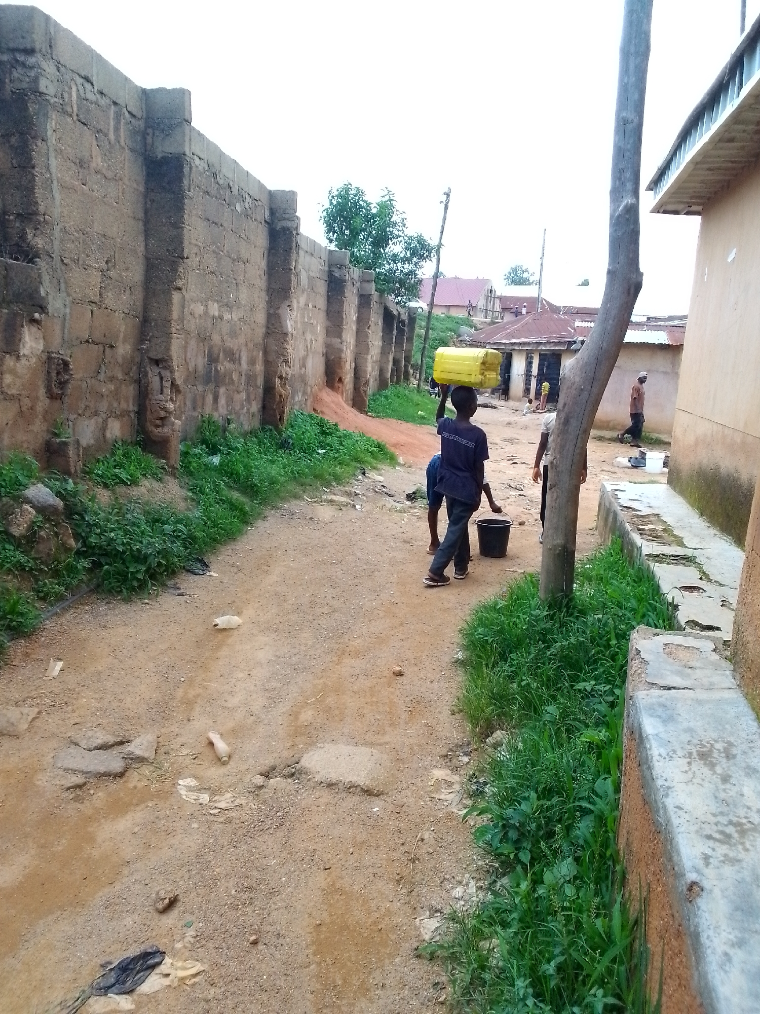 In an outdoor scene in a rural or urban fringe environment, a young person in a dark blue t-shirt and dark pants carries a yellow plastic container on their head. They are walking on a dirt path lined with a rough brick wall on the left and vegetation. Another young person is standing behind them, with a black bucket at their feet. Further in the background, several buildings with corrugated metal roofs are visible, along with other people. A large tree trunk stands prominently on the right side of the frame, near a concrete structure and more vegetation. The ground is uneven and dusty, with scattered debris including plastic bags and a discarded bottle. The overall impression is one of a utilitarian, developing community.