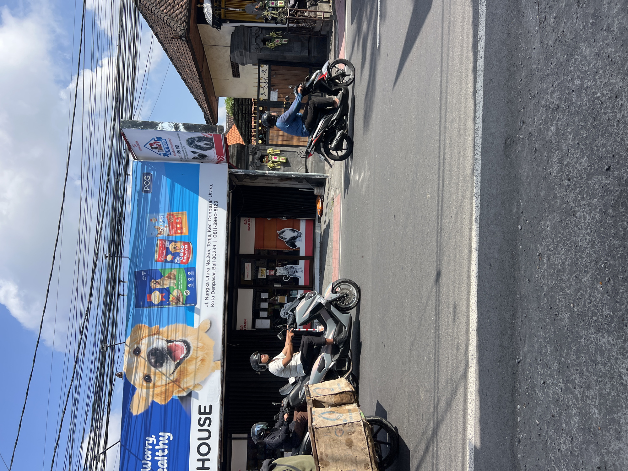 In Denpasar, Indonesia, on Jl. Nangka Utara No.265, Tonja, Kec. Denpasar Utara, two individuals are riding scooters on a paved street under a blue sky with scattered clouds. A large banner, possibly advertising a pet supply store, is displayed above the storefronts, featuring a smiling golden retriever and various pet food products. The banner also includes contact information: 0811-3960-8129. To the left of the frame, a wooden cart with two large bins is being pulled by a scooter. The architecture of the buildings suggests a tropical environment, with tiled roofs and decorative elements. Electrical wires are prominently strung across the top of the image.
