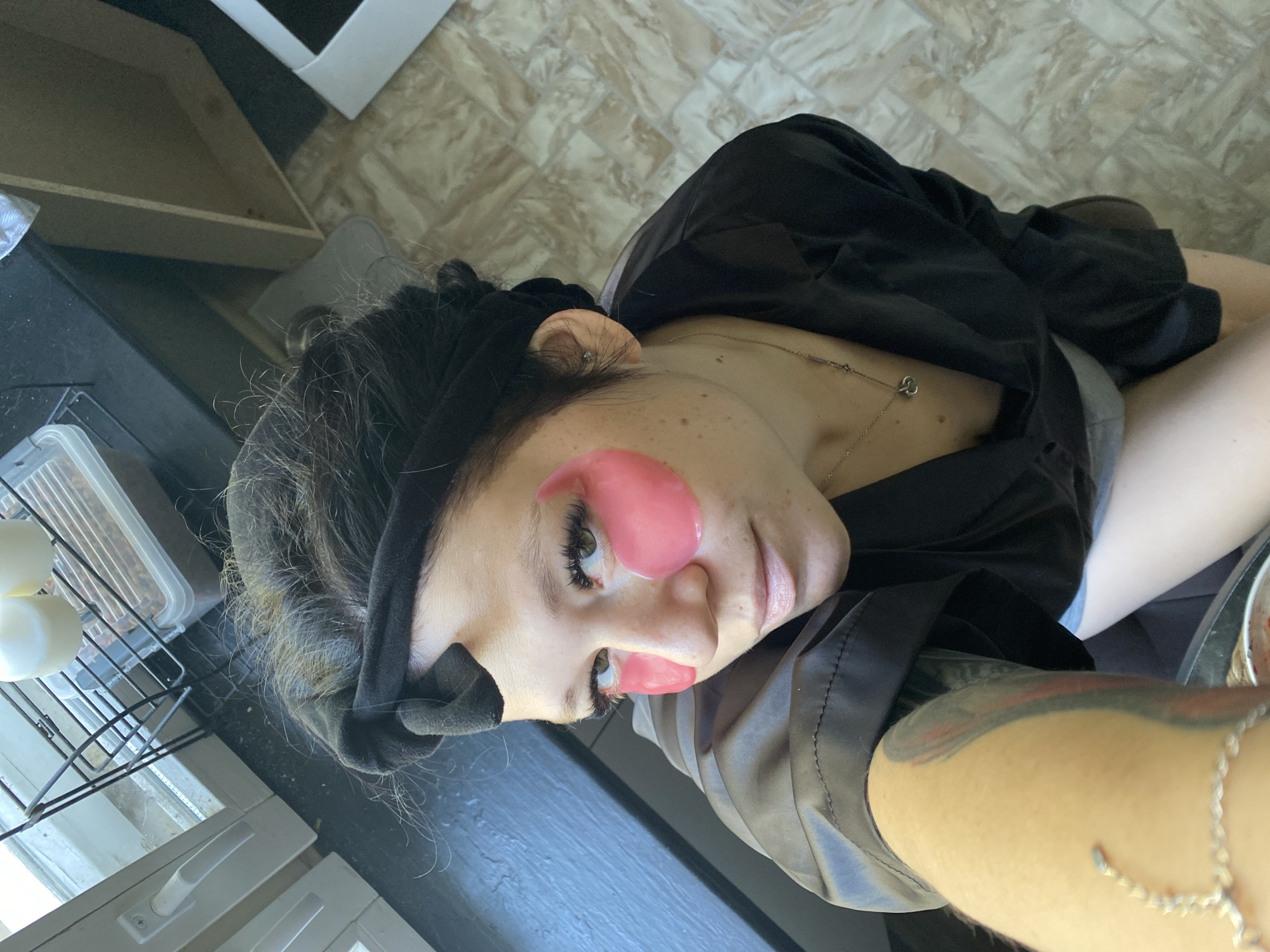 A woman with dark hair tied back with a black headband is shown from a high angle, looking up at the camera. She is wearing a black robe and has pink under-eye patches on her face. Her left arm, which is tattooed, is in the foreground, partially obscuring her body. The background suggests an indoor setting, possibly a kitchen or bathroom, with a window to the left and tiled flooring. There are no discernible landmarks that would place this image specifically in Chaikovsky, Russia.