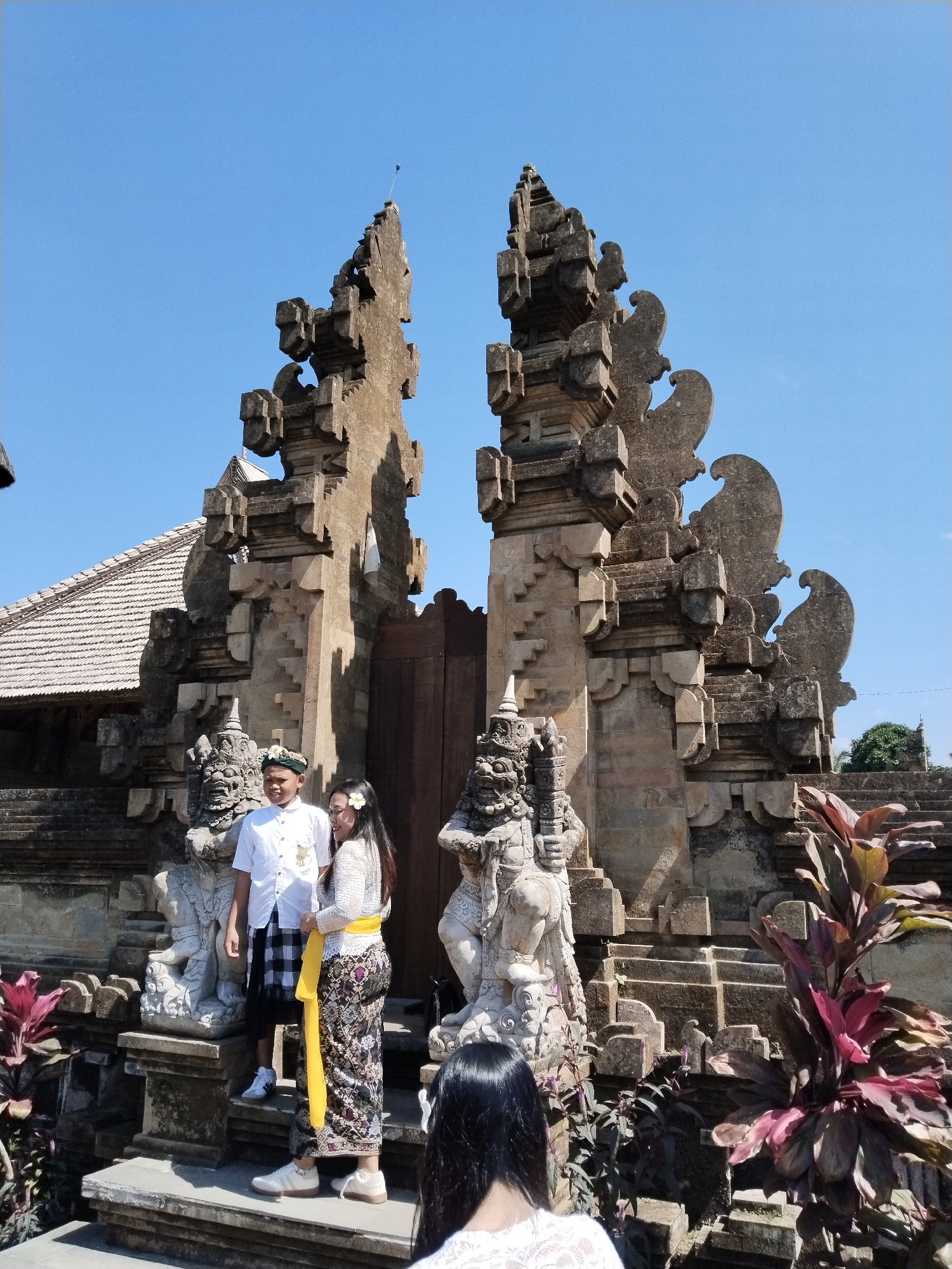 In Bangli, Indonesia, a young couple stands in traditional Balinese attire in front of a stone temple gate. The man wears a white shirt, a green headscarf, and a black and white checkered sarong. The woman is dressed in a white lace top and a patterned skirt with a yellow sash. They are positioned on a stone staircase leading up to the ornate temple entrance, which features intricate carvings and two guardian statues. The background shows a clear blue sky and the tiled roof of a building. The scene is captured during daylight, with sunlight illuminating the subjects and the architecture.