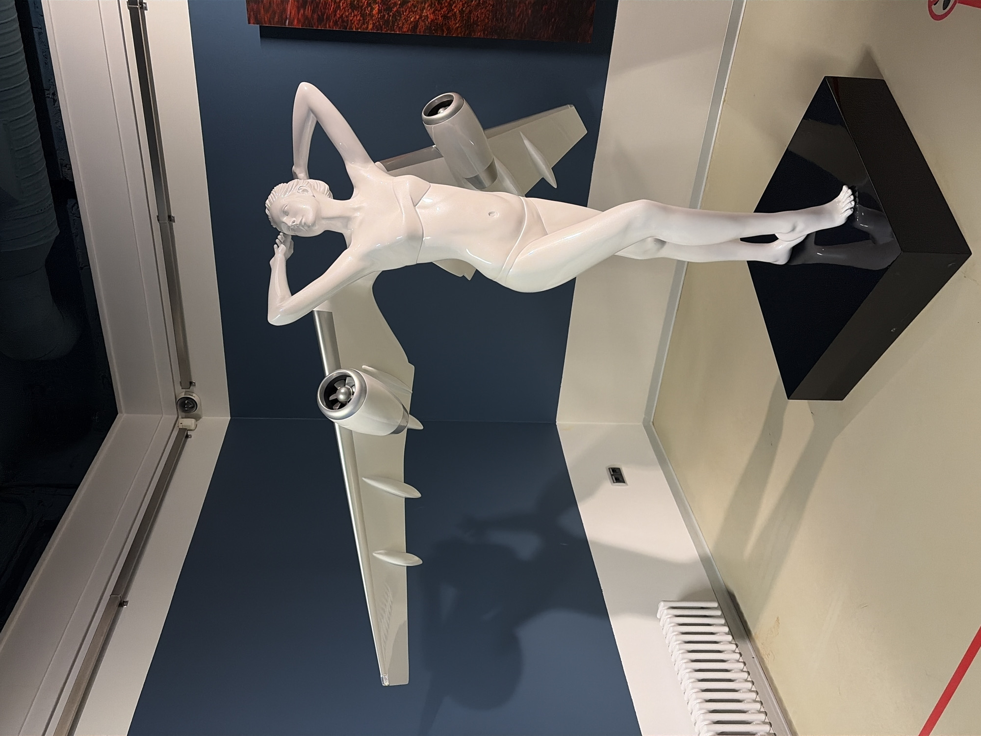 This image depicts a white mannequin resembling a female figure wearing a bikini. The mannequin is positioned in front of a dark blue wall, with its arms raised above its head. A white model airplane wing, complete with engines, is attached to the mannequin's back, extending outwards on both sides. The mannequin stands on a black rectangular pedestal. To the left of the mannequin, there is a white radiator against the wall. The floor is a light beige color, and a red line is visible on the floor in the lower left corner. In the background, on the right side of the frame, a portion of a red and black artwork is visible. The environment appears to be an indoor exhibition space. No specific location in Saint Petersburg, Russia is identified within the image.
