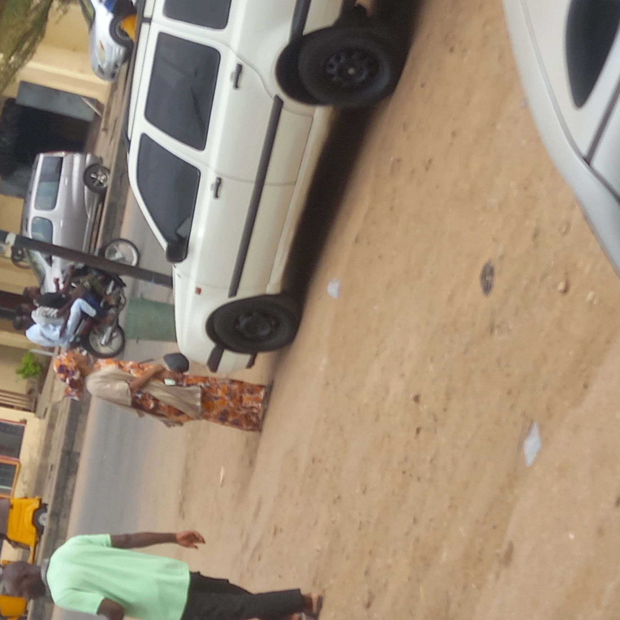 The scene depicts a street in what appears to be Dadi Riba, Nigeria. A white station wagon is parked near the curb, oriented at an angle to the road. A motorcycle with two riders is visible further up the street. A woman in traditional dress stands on the side of the road near the car. A man wearing a green shirt and dark pants is walking across the street in the foreground. A silver car is partially visible in the upper right of the frame. The road surface is dusty, and there is some trash on the ground.