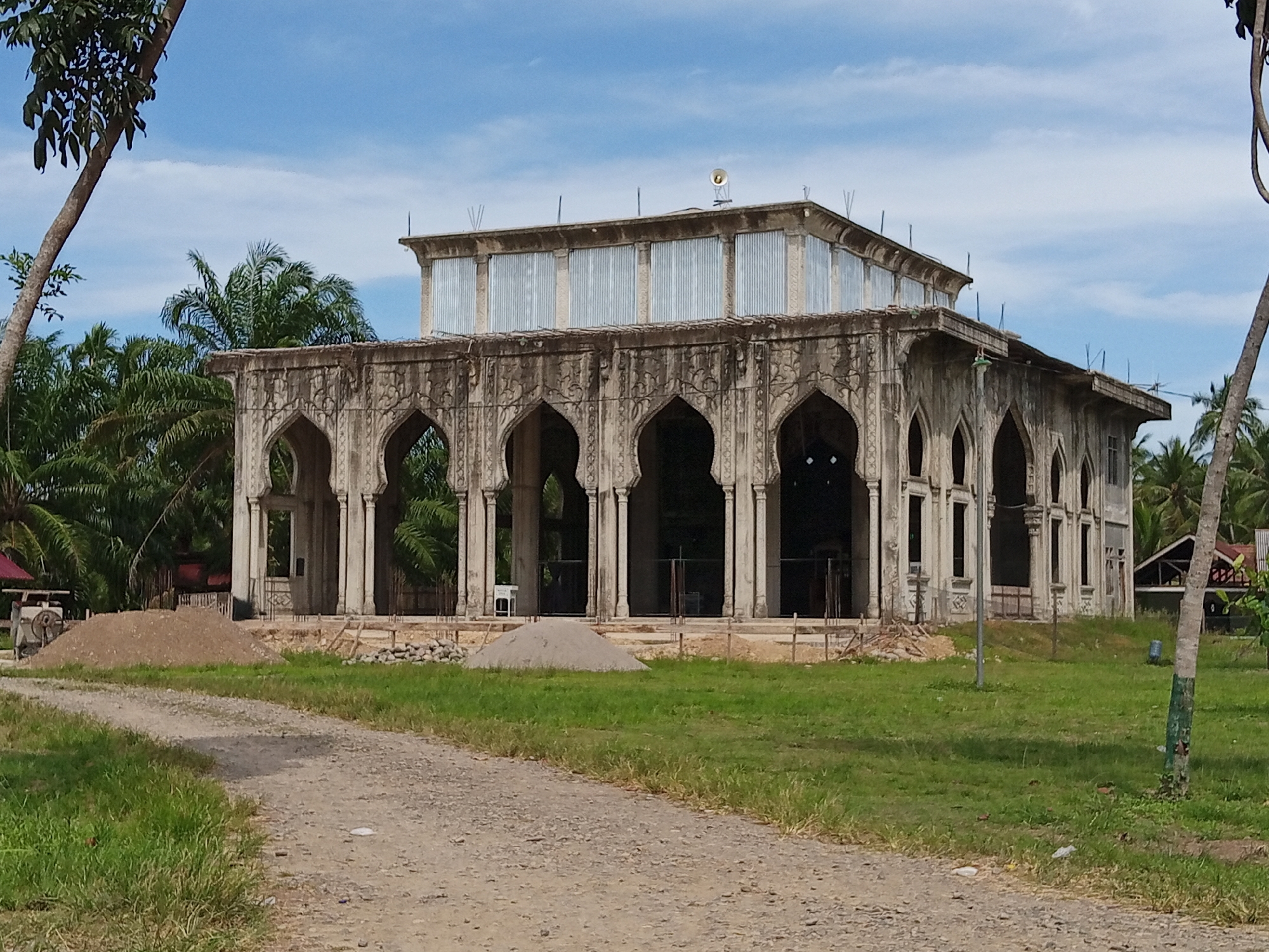 The image shows an incomplete building, likely a mosque, in Meunasahbeunot, Indonesia. The structure features a series of ornate arches and columns, but the upper level is constructed with corrugated metal panels. Construction materials are scattered around the base of the building, indicating ongoing work. A dirt road leads up to the building, and the surrounding landscape consists of grass and palm trees. The sky is partly cloudy.