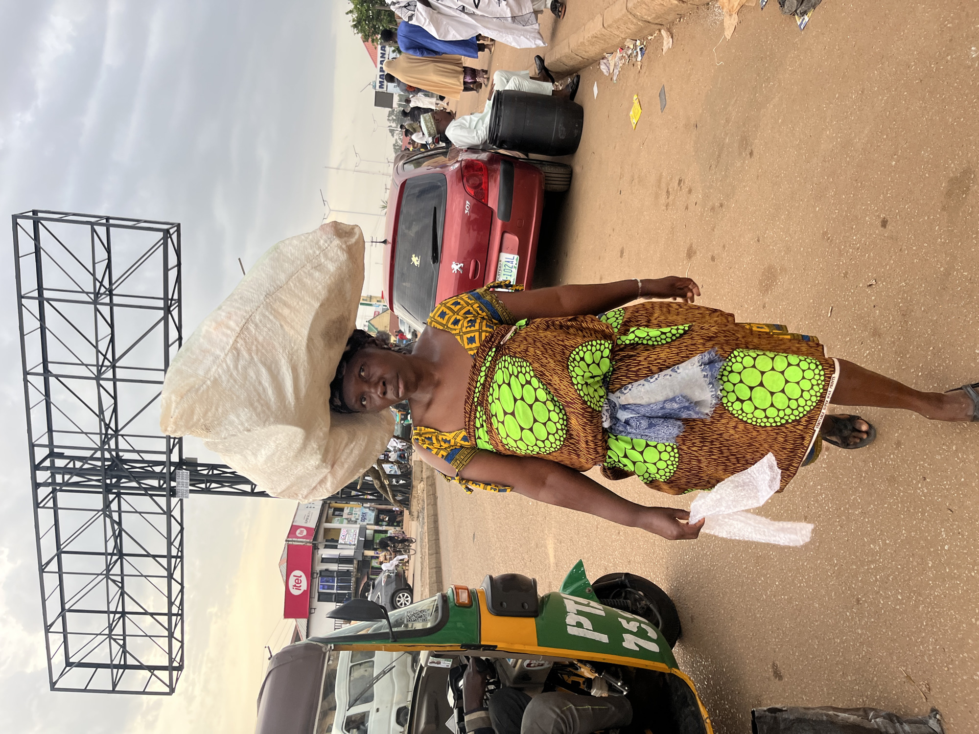 In Bukuru, Nigeria, a woman carries a large, light-colored sack on her head as she walks on a dirt road. She wears a vibrant dress with a brown base and a pattern of bright green circles. A blue and white scarf is tied around her waist. In her right hand, she holds a piece of white plastic. To her left, a green and yellow auto-rickshaw is parked. In the background, a red car is visible, along with other people and buildings, including a billboard overhead. The sky is overcast.