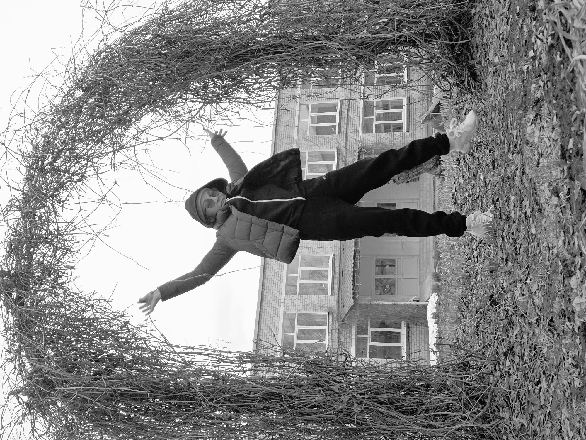 The black and white image, which appears to be rotated 90 degrees clockwise from its natural orientation, depicts a person striking a dynamic pose outdoors. The central figure stands with arms outstretched and legs spread wide, suggesting a moment of balance, joy, or performance. They are dressed in casual attire, including a hooded jacket or vest, dark pants, and light-colored sneakers. Round, dark sunglasses obscure their eyes.
The person is framed by an archway of dense, intertwined bare branches, likely vines, which dominate the foreground and sides of the composition, creating a rustic, natural frame. To the right, a wall or structure is covered in thick foliage, contrasting with the barren branches and hinting at a different type of plant life. In the background, a multi-story brick building with numerous rectangular windows provides an architectural backdrop, suggesting an urban, institutional, or residential setting. The sky is bright, indicating it is daytime, though the lack of strong shadows could mean it's an overcast day or early/late sunlight. The bare branches suggest the season is late autumn or winter. There is no visible text in the image. The overall impression is one of playful interaction with the environment.