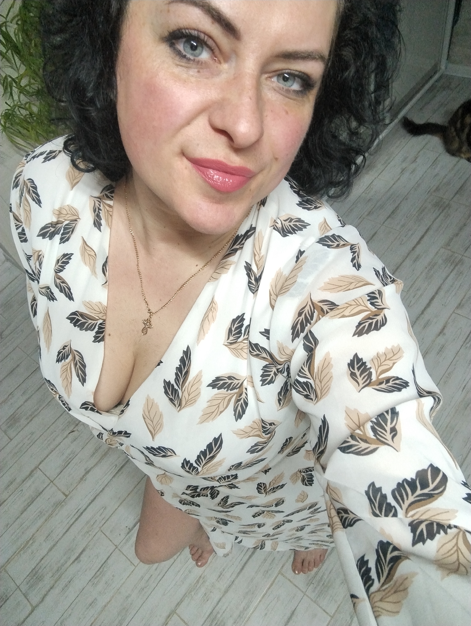 A high-angle indoor selfie captures a woman from the chest up, looking directly at the camera with a slight smile. She has dark, curly hair and striking blue eyes. Her face shows light makeup, including pink lipstick and blush, and some freckles are visible. She is wearing a white dress with a pattern of dark brown and light brown leaves. The dress features a V-neckline, revealing some cleavage, and she wears a delicate gold chain necklace with a small cross pendant. Her bare feet, with painted toenails, are visible at the bottom of the frame, resting on light-colored tile or laminate flooring. In the background, to the left, green foliage from a houseplant is partially visible. To the right, a domestic cat with tabby markings is partially seen on the floor. The lighting suggests an indoor setting, likely a home, in Grishanki, Russia. There is no visible text in the image.
