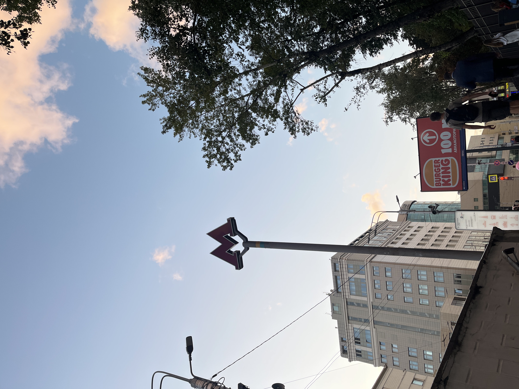 The image captures an outdoor urban scene in Moscow, Russia, under a pale blue sky with scattered, light pink clouds, suggesting late afternoon or early evening. In the foreground, a dark, textured wall runs horizontally. Above this, a tall, grey pole supporting a prominent red "M" logo, characteristic of the Moscow Metro, stands out against the sky. To the left of the metro sign, a streetlamp with a modern design is visible, along with a network of overhead wires. In the mid-ground, a large, multi-story building with a light grey facade and numerous windows is prominent on the left. To the right of the metro sign, a billboard advertising Burger King with the text "100 M" and an upward arrow is displayed. A group of people are walking away from the camera towards the billboard, with one individual wearing a backpack. Further to the right, a dense canopy of green trees lines a black metal fence. The overall impression is of a bustling city street corner, blending urban infrastructure with elements of nature.