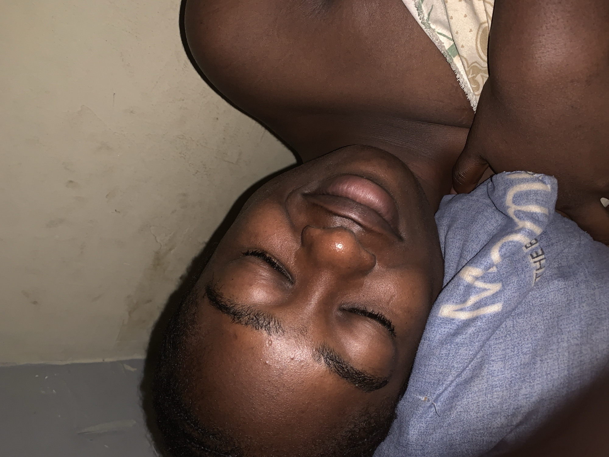 This close-up, rotated image features a person with dark skin sleeping or resting. The subject lies on their side, with eyes closed and lips slightly parted, conveying a sense of peaceful slumber. They have short, dark hair and visible stubble on their face. Their head is positioned on a blue, denim-like fabric, which appears to be a pillow or a soft covering. White text is visible on this blue fabric; "THEB" is clearly discernible, along with a partial, stylized letter that resembles an "M" or "W."
The upper part of the person's torso is visible, partially covered by a light-colored sheet or blanket with a subtle pattern. One arm or hand is tucked near their neck or shoulder. The background reveals a simple, light-colored wall with visible texture, some discoloration, and minor imperfections, suggesting an indoor setting. The lighting in the image is dim, indicating either nighttime or a poorly lit room. The scene evokes quiet and rest, with the person appearing undisturbed in their sleep. No specific activity beyond sleeping is depicted.