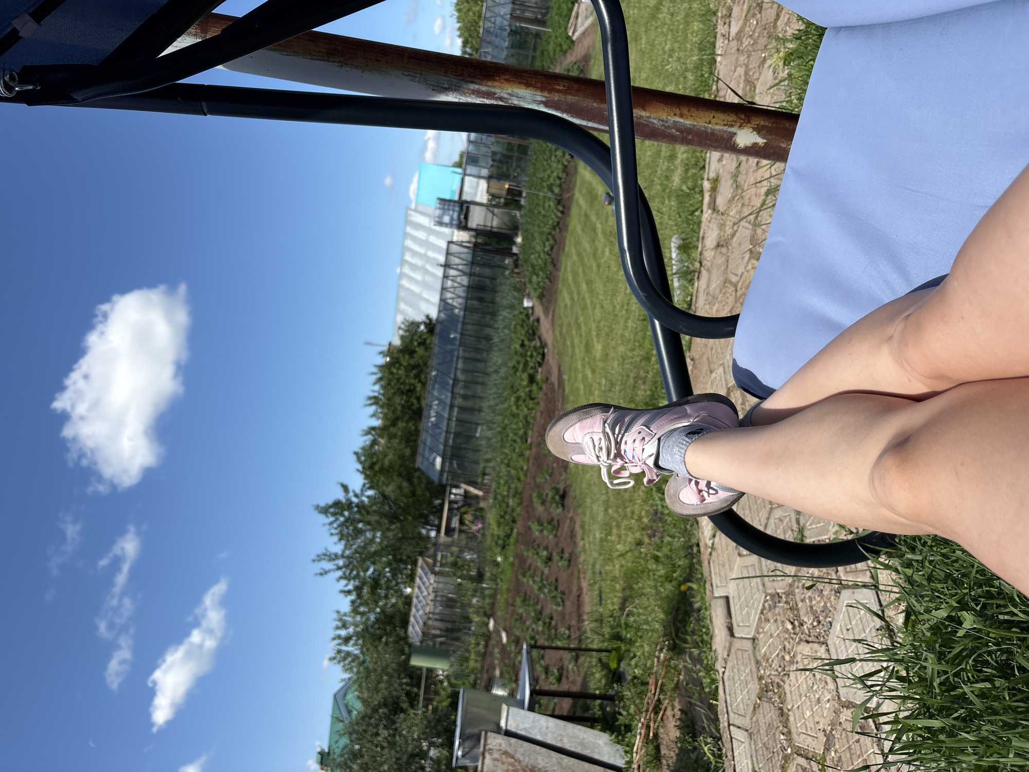 The image depicts a person's legs and feet resting on a blue cushioned swing seat outdoors. The person is wearing pink sneakers and grey socks. The swing is set within a garden area. In the background, there is a tilled garden bed with rows of plants, several greenhouses, and trees. The sky is blue with scattered white clouds. The location is identified as Прикамский, Russia, suggesting a rural or suburban setting. The scene appears to be a sunny day.