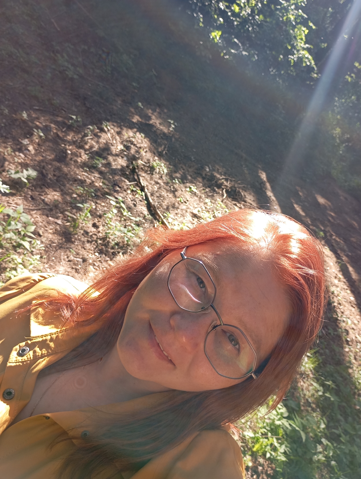 A close-up selfie-style portrait captures a young adult female outdoors in Chaikovsky, Russia. She has striking reddish-auburn hair and fair skin, illuminated by bright sunlight. She wears distinctive glasses with thin, dark, somewhat octagonal frames. Her expression is a gentle, pleasant smile. She is dressed in a mustard yellow button-up shirt or light jacket, and a delicate silver-colored necklace is visible around her neck, along with a faint, light-colored circular mark or pattern on her upper chest.
The image is taken from a slightly low angle, looking upwards. Strong sunlight streams from the upper right, creating noticeable lens flares and casting dappled light on the background. The setting appears to be a natural, wooded area, possibly a forest or park. The ground is visible behind her, covered with brown dirt, scattered leaves, and patches of green undergrowth and small plants. The bright sun suggests a clear day, likely morning or late afternoon. No visible text is present in the image.