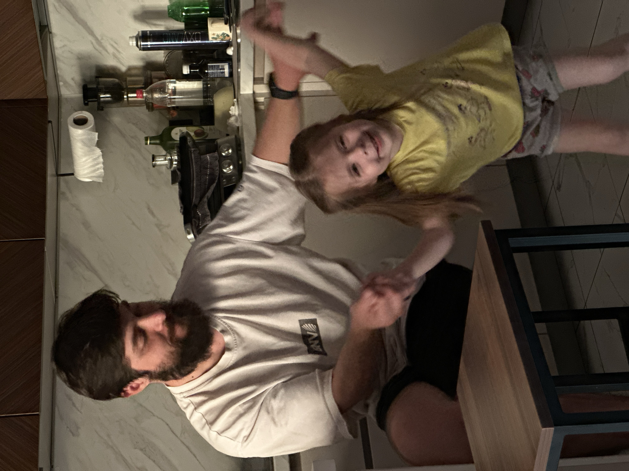 A man with a beard, wearing a white t-shirt and black shorts, is interacting with a young girl with long brown hair, wearing a yellow t-shirt and patterned shorts. They appear to be holding hands and swinging the girl around. The location appears to be indoors, possibly a kitchen or bar area, given the presence of bottles on a shelf and a roll of paper towels. A table with a wooden top and black metal frame is also visible. The precise location in Kuda, Russia is indeterminable from the image.