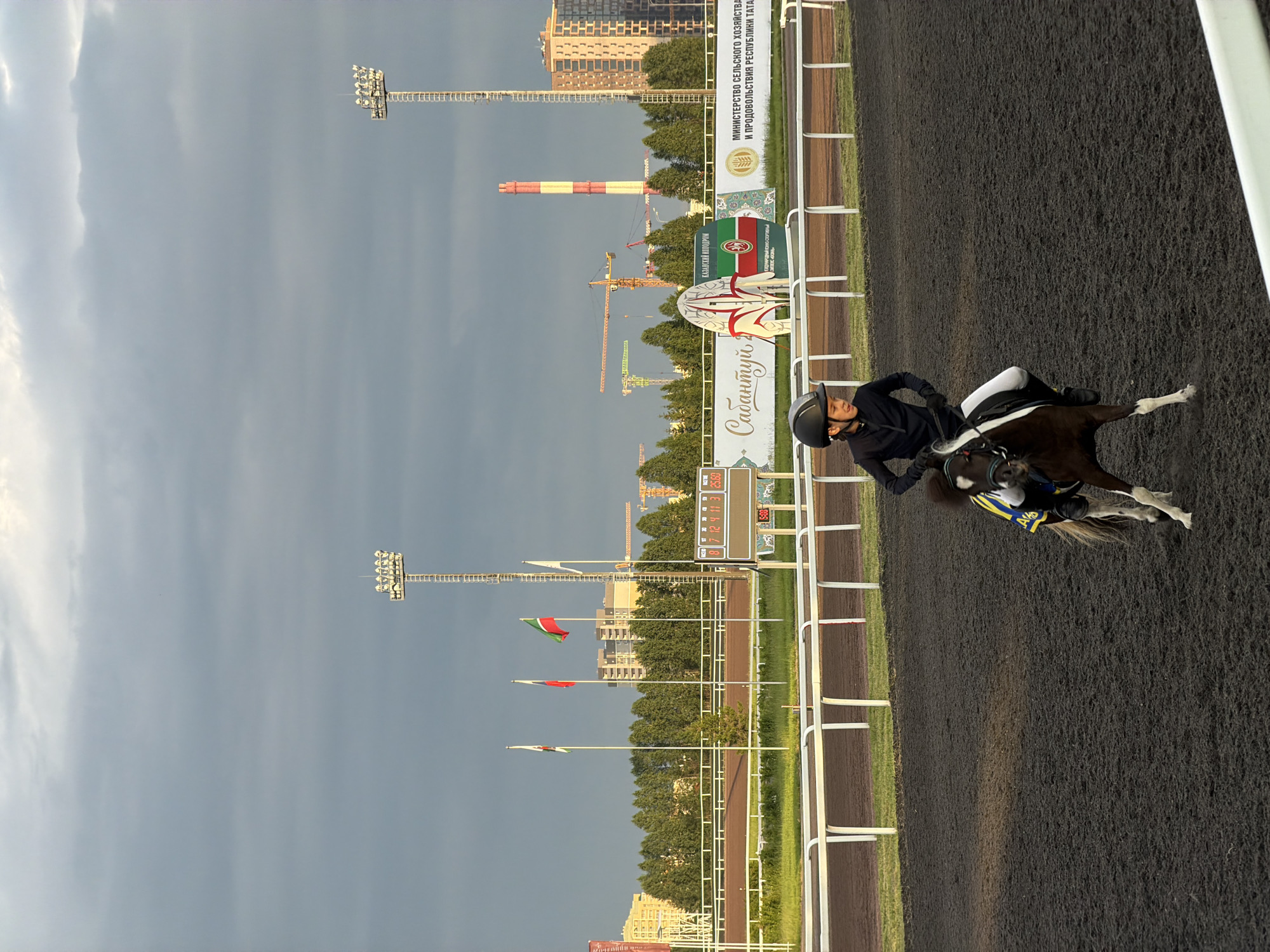 A young boy in a black riding outfit and helmet is riding a miniature horse on a dirt race track. The horse is dark brown with white markings on its legs and face, and it is wearing a yellow and blue blanket. The boy is looking forward and appears to be concentrating on riding. In the background, there is a white fence lining the track, followed by a row of trees, and beyond that, several tall buildings and industrial structures, including cranes and a tall red and white striped chimney. Two large floodlights are visible in the sky. The sky is overcast with dramatic clouds. A digital scoreboard displaying numbers is also visible near the fence. Flags are flying on poles in front of the trees. The scene appears to be a horse racing or equestrian event taking place in an urban environment during the day.