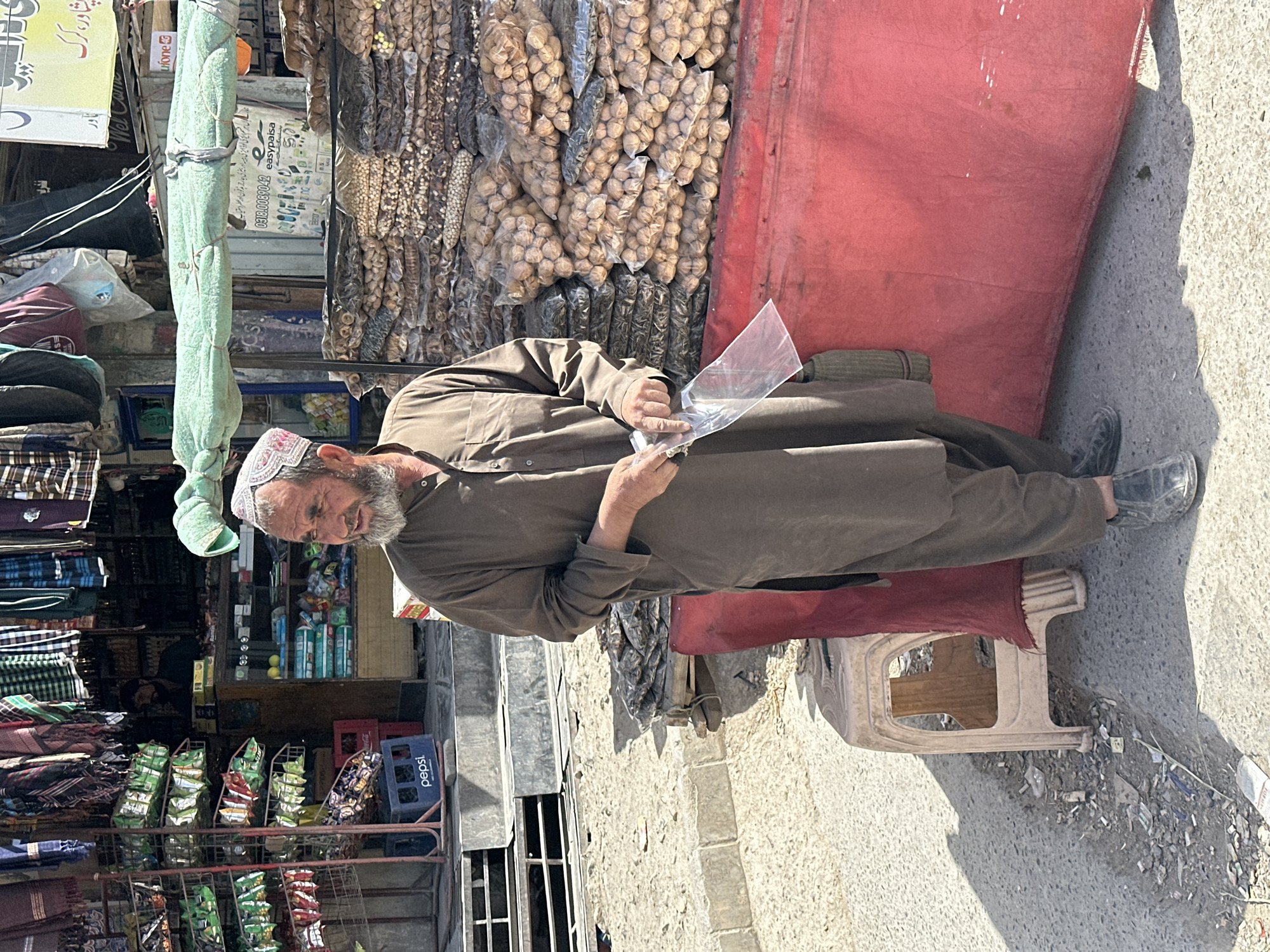 The image captures a candid moment in an outdoor market setting, likely in Pakistan, given the visible text. The central figure is an older man with a grey beard and a white, embroidered cap (koofi). He is dressed in a traditional long, loose, brown garment. The man appears to be inspecting a clear plastic bag in his hands, possibly containing a product for sale or purchase.
He is positioned beside a makeshift stall, characterized by a large red tarp or fabric that serves as a backdrop. Hanging abundantly from the stall are numerous clear plastic bags filled with various dried goods, including what appear to be walnuts, chickpeas, dried corn/maize, and sunflower seeds, all neatly packaged for individual sale.
To the left of the man, the scene reveals more of the market. There are shelves stacked with rolls of colorful fabrics or textiles. Further into the background, another shop or section of the stall displays various packaged snacks like chips and biscuits arranged on metal racks. A blue Pepsi crate is also visible. The ground is concrete or asphalt, showing signs of wear and some light litter, and a light-colored plastic stool stands empty nearby.
The bright sunlight and sharp shadows indicate it is daytime, likely a clear and sunny day. Visible text on signs includes "easypaisa" and "Ufone" (Pakistani mobile financial and telecommunication services, respectively), a phone number starting with "031", and partially visible Urdu script that includes "پشاور" (Peshawar) on a yellow sign, strongly indicating a location in Pakistan. The man's expression is neutral, seemingly focused on the item in his hands.