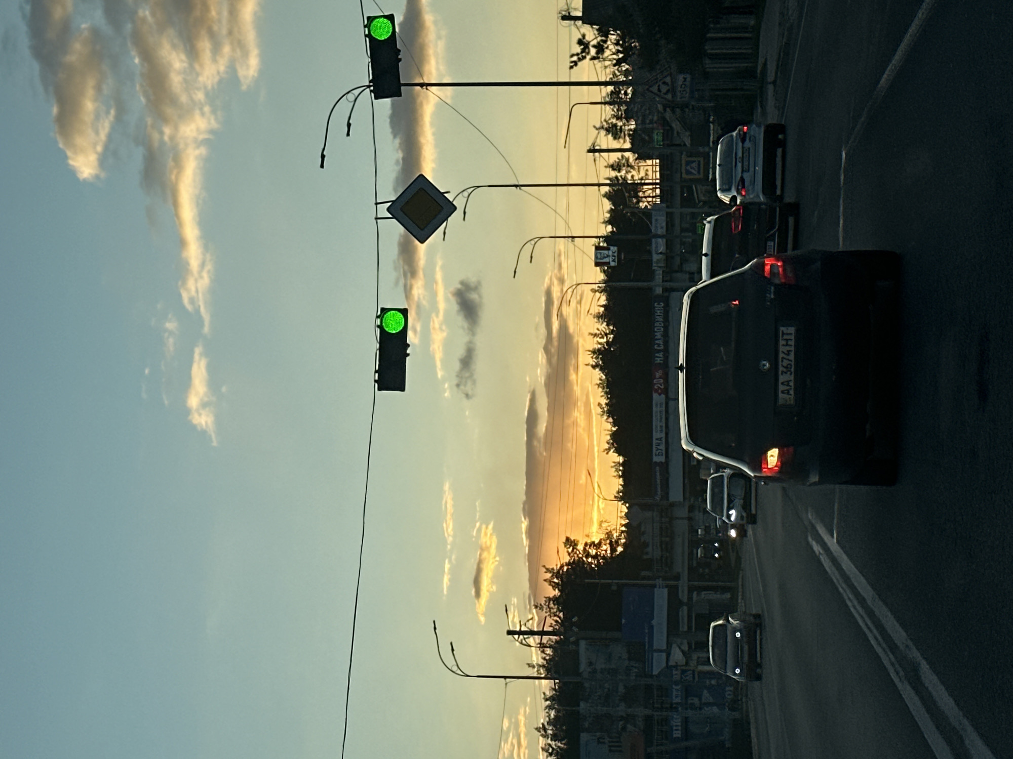 The image depicts an evening road scene at sunset in Hostomel, Ukraine. The sky presents a gradient of colors, transitioning from a light blue at the top to a warm orange and yellow glow at the horizon, where the sun is setting. Scattered, elongated clouds are illuminated by the golden light.
On the left side of the frame, two green traffic lights are visible on poles, indicating that traffic can proceed. Between them, a yellow diamond-shaped road sign with a white border, signifying a priority road, is prominent. Several streetlights with curved arms are positioned along the road.
The multi-lane asphalt road is populated with vehicles. The dark-colored car closest to the viewer displays a Ukrainian license plate: "AA 3674 HT". Further along the road, a white car and other dark vehicles are visible, some with their headlights or taillights illuminated.
In the background, behind a line of trees, various urban elements are discernible. A billboard or sign features Cyrillic text, including "БУЧА" (Bucha), "-20%", and "НА САМОВИВІС" (for self-pickup), along with a "KFC" logo. Other road signs, such as one marked "150M" and a small triangular warning sign, are also present. The overall scene captures the typical ebb and flow of evening traffic under a dramatic sunset sky.