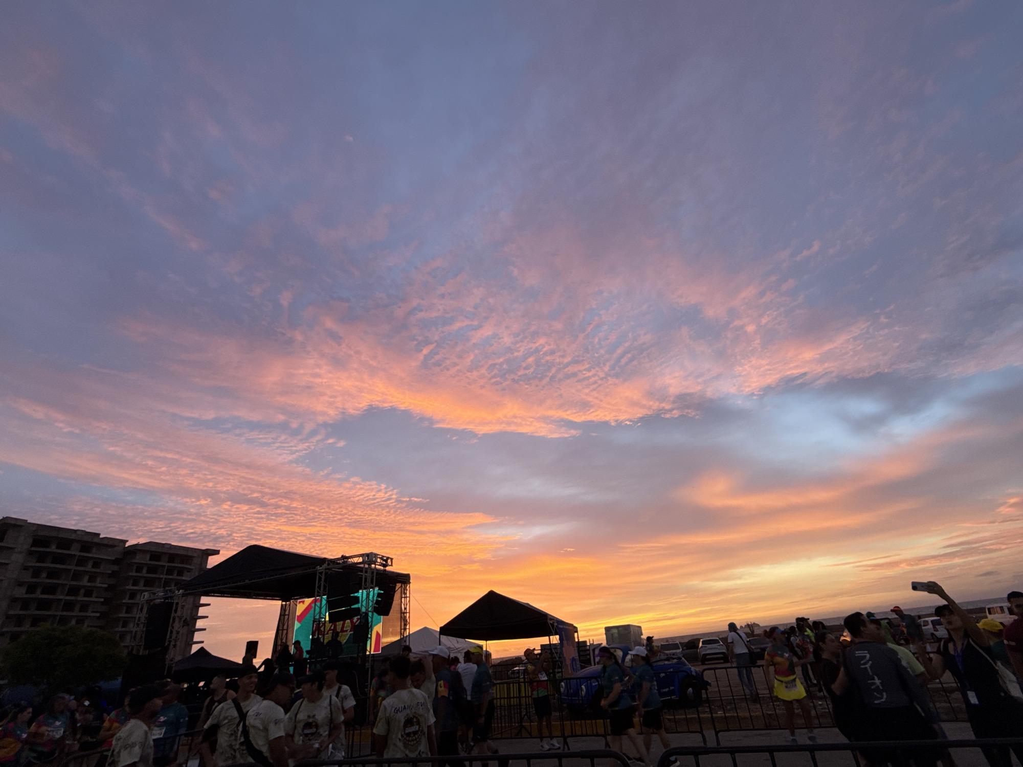 The image depicts an outdoor event at sunset in Porlamar, Venezuela. The sky dominates the upper two-thirds of the frame, showcasing a vibrant sunset gradient ranging from deep orange and red at the horizon to pinks and purples higher up, interspersed with numerous textured altocumulus or cirrocumulus clouds reflecting these colors.
In the midground, a large, dark-canopied stage structure with exposed trussing and two prominent LED screens displaying colorful, abstract graphics (green, yellow, red) is central. To the immediate left of the stage stands a multi-story, unfinished or abandoned concrete building with numerous empty window frames. To the right of the stage, a smaller, dark pyramidal tent is visible.
The foreground and midground are densely populated with a crowd of individuals, primarily adults, many obscured or blurred. Black metal crowd control barriers separate the viewer's immediate perspective from the gathering. Visible individuals include a person in a light t-shirt with "GUAMPBE" text, another in a dark t-shirt with Japanese characters "ラーメン" (Ramen), and a person in a yellow skirt or shorts with a visible race bib. Further right, an individual is holding a smartphone aloft, seemingly capturing the scene. In the background, behind the crowd and barriers, several vehicles, including at least two blue jeeps, are parked. People are standing and congregating, suggesting attendance at a public gathering or performance.