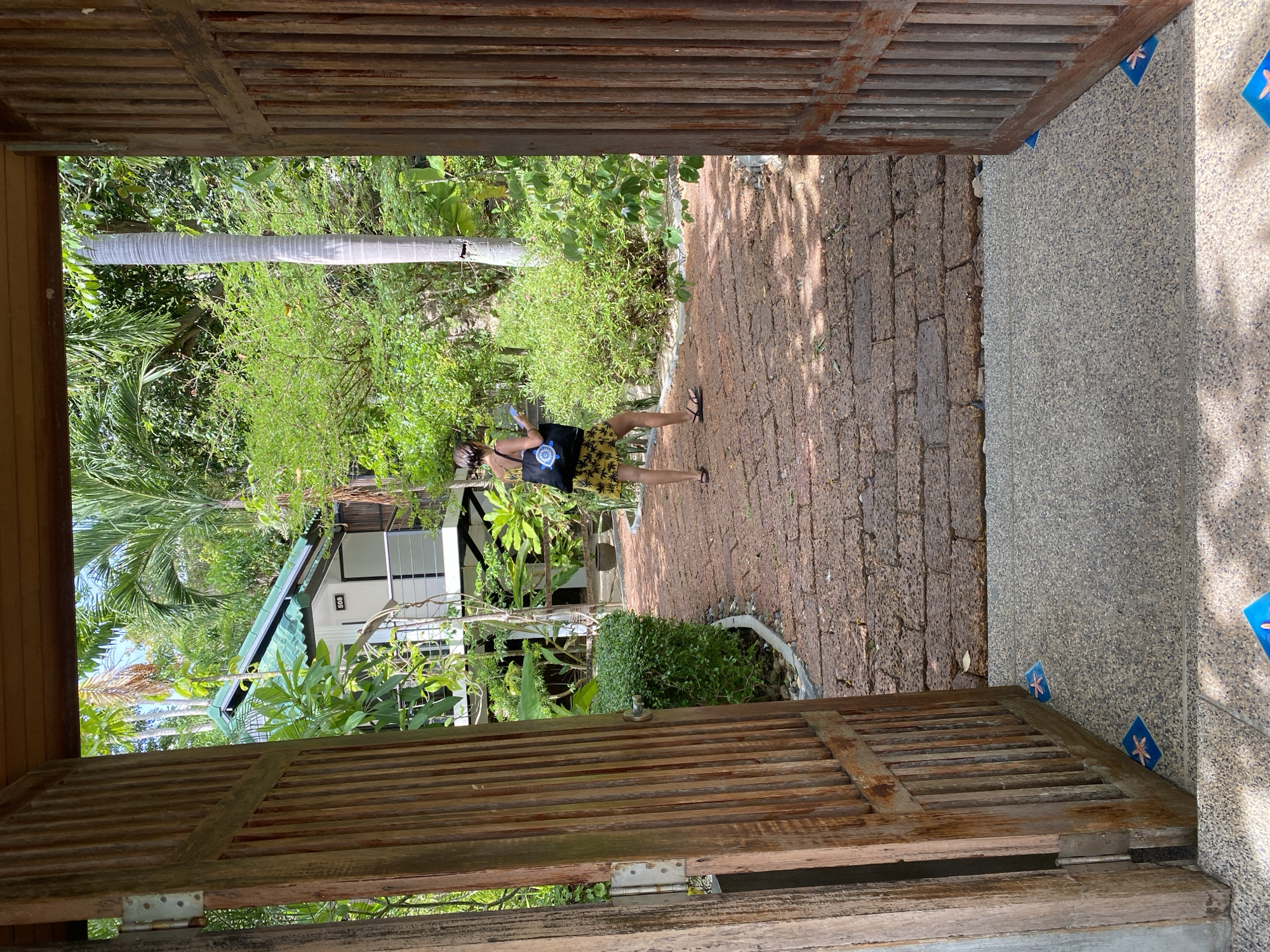 A person is standing on a brick pathway, holding a phone in their hand. They are wearing a yellow dress with a black and white pattern and black sandals. A black tote bag with a white turtle design hangs from their shoulder.
The scene appears to be outdoors in a tropical location, with lush green vegetation and palm trees visible in the background. A building with a green roof and white walls can be seen amongst the foliage. The pathway is made of red bricks, and there is a textured grey surface next to it, possibly a sidewalk or building exterior, with small blue tiles featuring a pink starfish design.
The image is taken from a slightly elevated perspective, looking through an open wooden doorway with slatted doors. The lighting suggests it is daytime, likely sunny, as indicated by the shadows cast on the ground. The overall atmosphere seems relaxed and casual, possibly a resort or a residential area in a warm climate. The location is identified as Koh Phangan, Thailand.