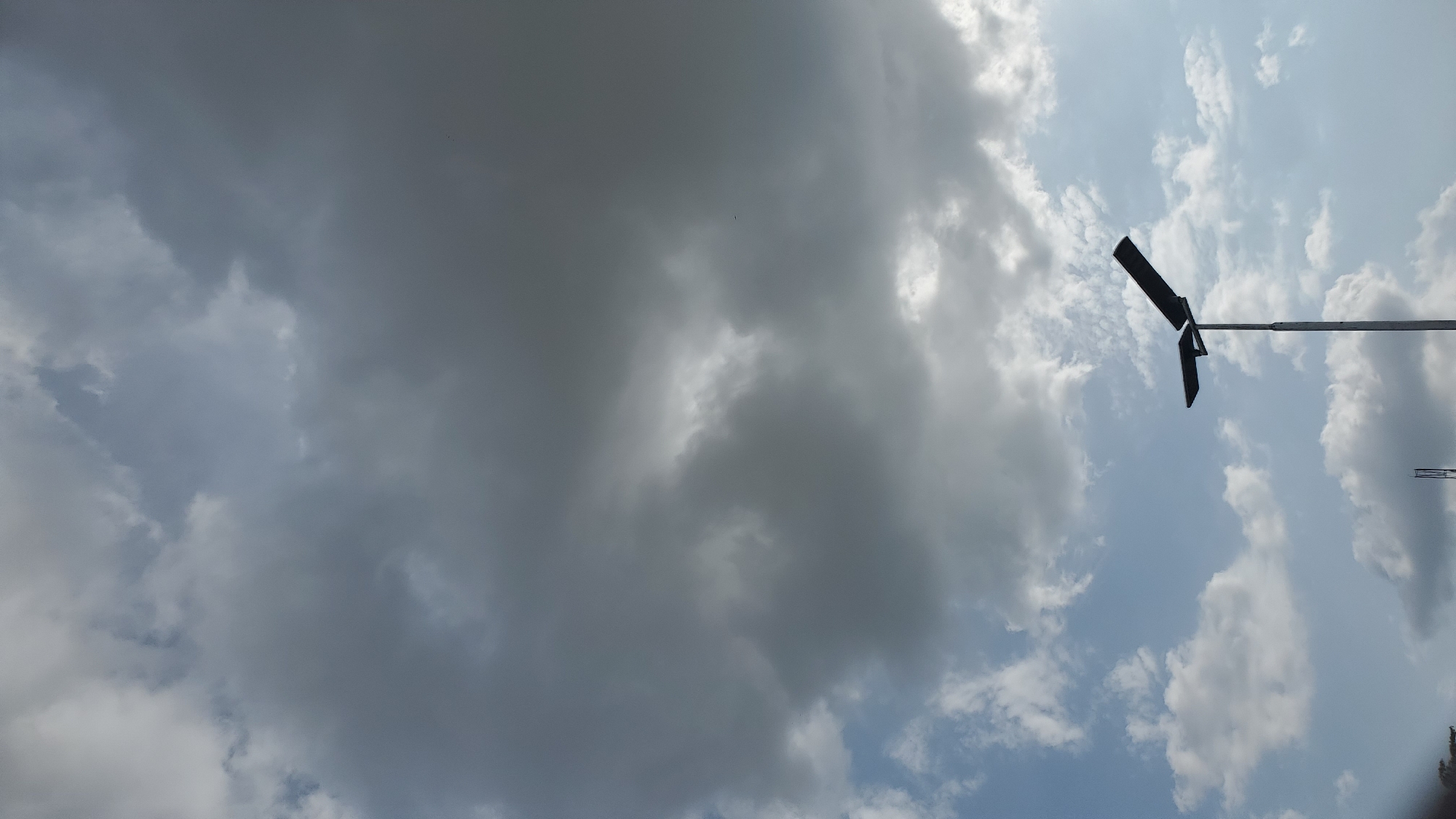 A street lamp with two solar panels stands silhouetted against a cloudy sky in Jalingo, Nigeria. The sky is a mix of dark, overcast clouds and patches of brighter, lighter clouds, with some hints of blue sky peeking through. The street lamp is tall and slender, with a dark pole and two dark, rectangular solar panels attached to the top, angled to capture sunlight. The overall impression is one of an overcast day, with the artificial light source of the street lamp visible against the natural backdrop of the sky.