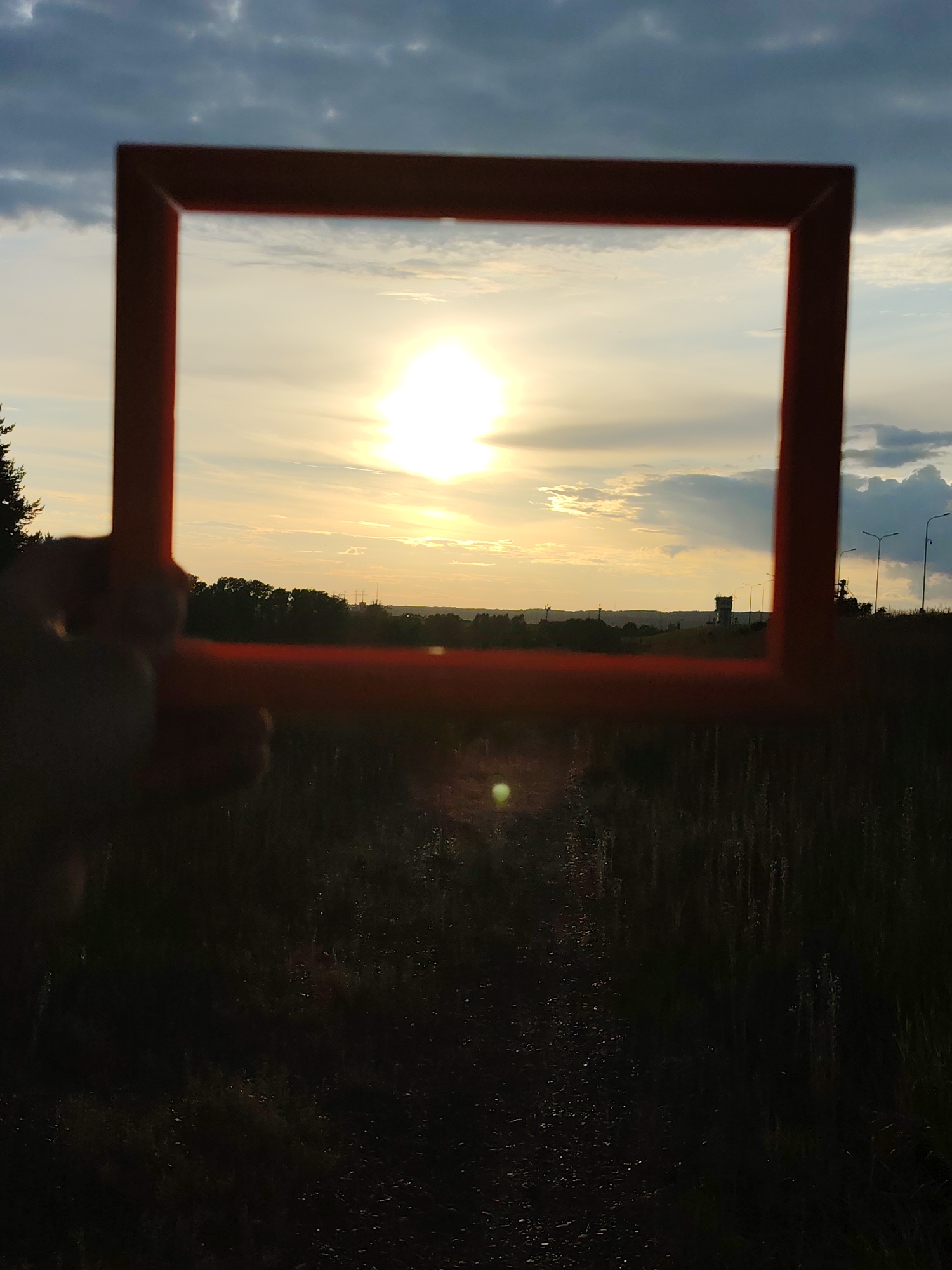 An orange frame is held in the foreground, obscuring the lower portion of the image. Through the frame, a sunset is visible over a flat landscape. The sky is filled with a mix of dark, cloudy formations and lighter, scattered clouds, illuminated by a bright, hazy sun. A line of dark trees forms a silhouette along the horizon. In the mid-distance, silhouetted against the sky, are several structures, including what appears to be a tall tower or industrial chimney on the right. Further to the right, streetlights are visible. The foreground consists of a dark, out-of-focus field with tall grass. The image is taken in the city of Chaikovsky, Russia.