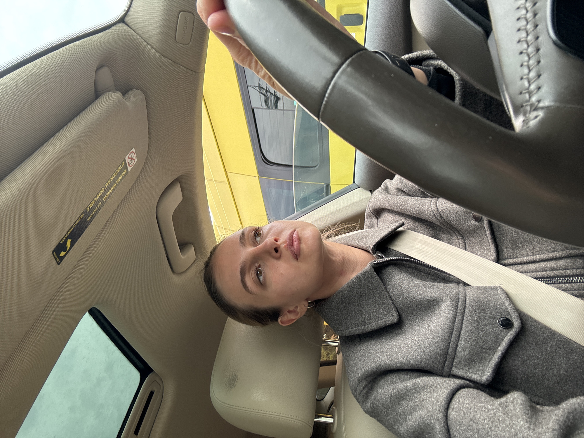 A woman is depicted from a high-angle, close-up perspective inside a car, likely taking a selfie. She is seated in what appears to be the driver's seat, looking upwards and slightly to her left with a neutral, contemplative expression. Her hair is pulled back, and she is wearing a grey wool or felt jacket and a light-colored seatbelt across her chest.
The car's interior features light beige upholstery, including the headliner, sun visor, and seat. A dark grey steering wheel is prominent in the foreground, partially obscuring the view through the front windshield and passenger-side window. Through the windows, a bright yellow structure or vehicle is distinctly visible outside, creating a vibrant color contrast with the car's interior. The sky visible through the sunroof or upper part of the window appears overcast.
Visible text includes "AIRBAG" on the car's A-pillar and a safety label on the sun visor that reads "AIR BAG WARNING / ATTENTION SAC GONFLABLE" in both English and French. The scene suggests a daytime setting, possibly during a stop or a period of waiting, within a vehicle in Tbilisi, Georgia.