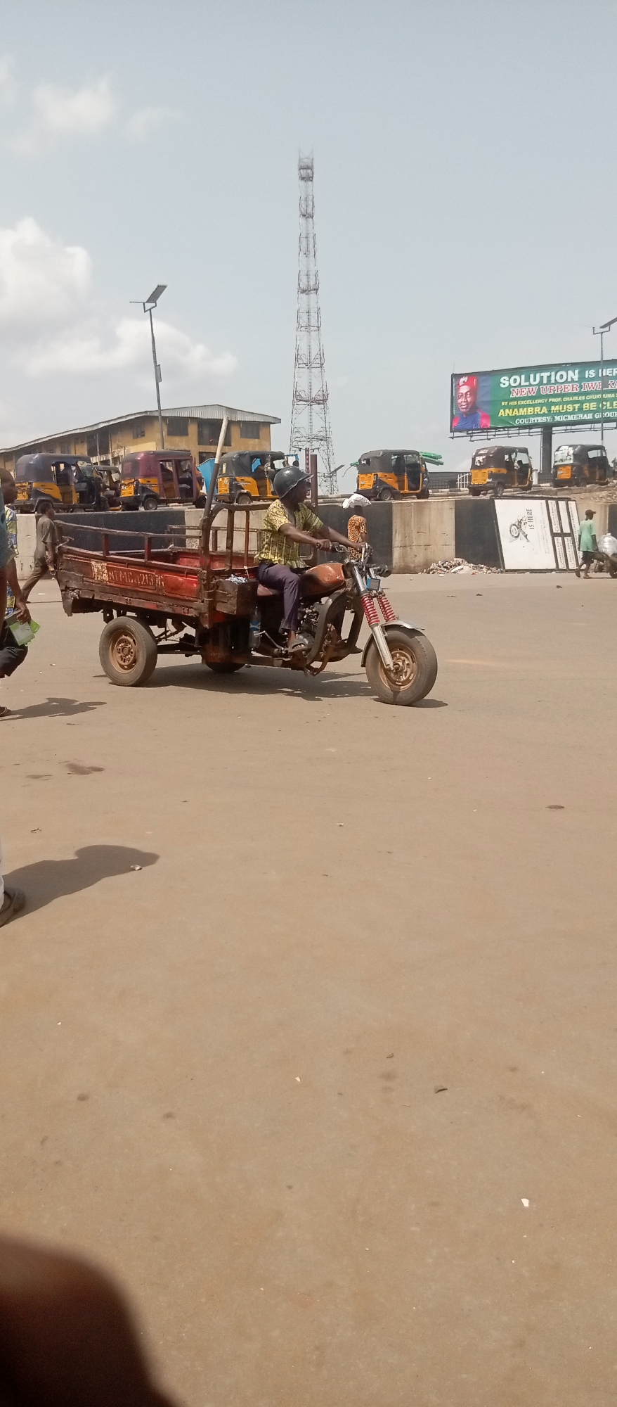 The image captures a scene in Onitsha, Nigeria, featuring a man operating a rusty, three-wheeled cargo motorcycle. The vehicle is laden with various objects and has lettering that reads "MTMG C21376" on its side. The man wears a helmet and is dressed in a patterned shirt and dark pants.
In the background, there are several yellow auto-rickshaws parked along a concrete barrier. A large billboard is visible, displaying a political advertisement that reads "SOLUTION IS HERE," with the text "NEW UPPER IWE KA" and "ANAMBRA MUST BE CLEAN," suggesting it may be related to elections. A tall communications tower stands in the distance. The overall environment appears to be a busy intersection.