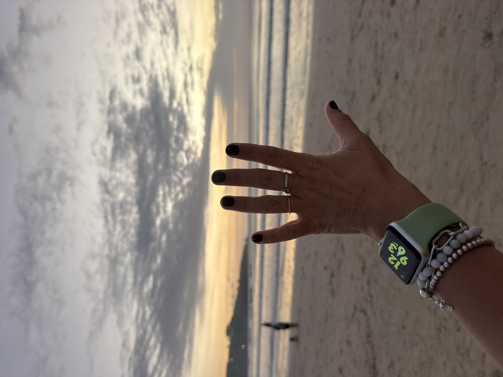 A medium shot captures a person's left hand with black nail polish and rings raised against a blurred backdrop of Kuta Selatan, Indonesia, during twilight. The hand is extended, palm facing the viewer, with fingers splayed. A smartwatch with a green band displays a colorful clock face on the wrist, adorned with layered bracelets. In the background, the calm ocean meets the sandy beach, with gentle waves rolling in. The sky is a mix of grey clouds and the warm hues of sunset, casting a soft light over the scene. Two indistinct figures and a dog are visible on the beach in the distance, adding a sense of scale and activity to the otherwise serene environment. The overall impression is one of tranquility and reflection at the end of the day.