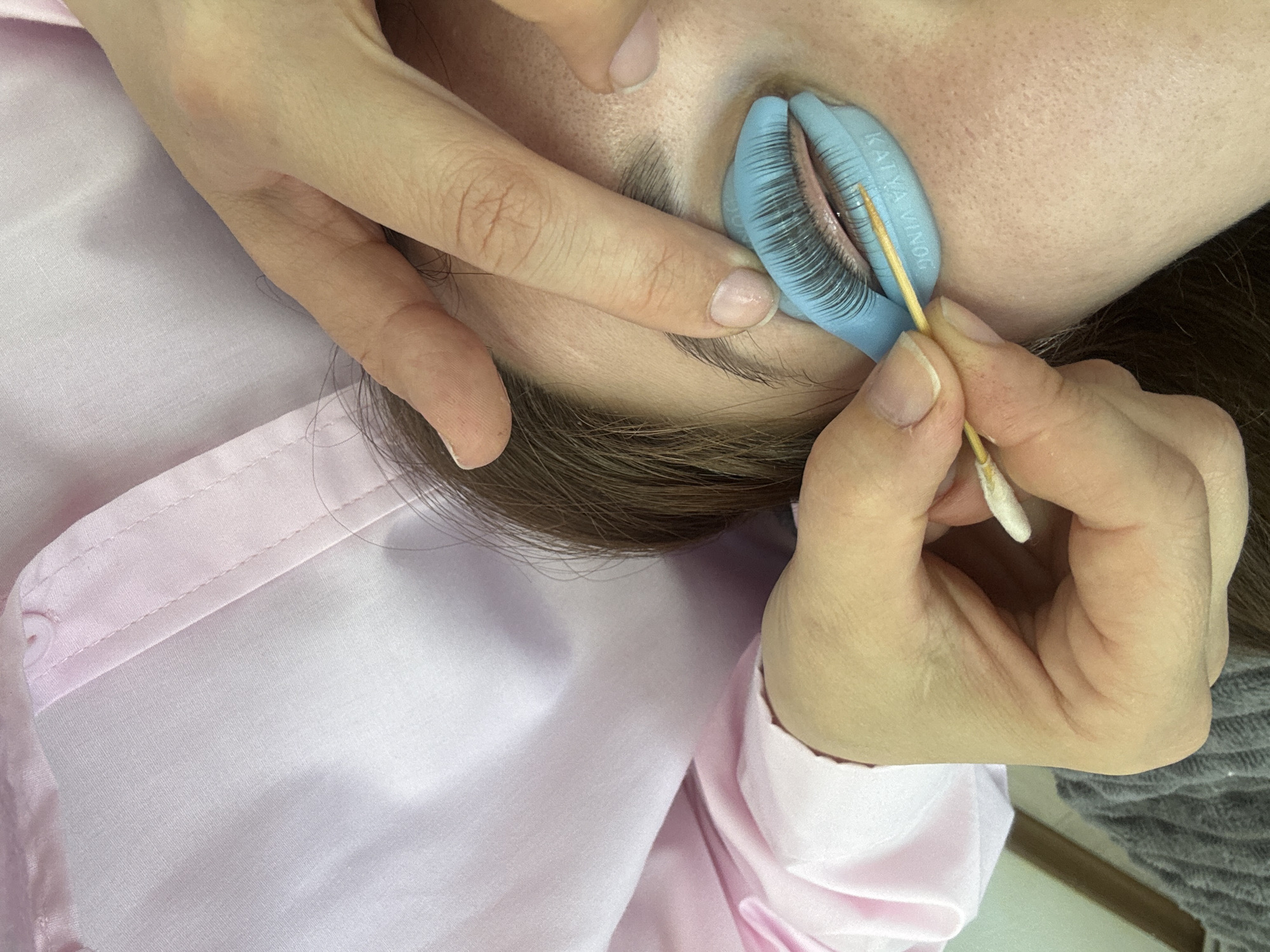 The image shows a close-up of a person's eye during a lash lift procedure. A blue silicone shield is placed under the upper lashes, and a wooden stick is used to separate and lift the lashes. The person's face is partially visible, with their eye closed and a pink shirt visible on their upper body. The scene appears to be a beauty salon or a home setting where such treatments are performed. The image does not provide any information about the location in the city of Chaikovsky, Russia.
