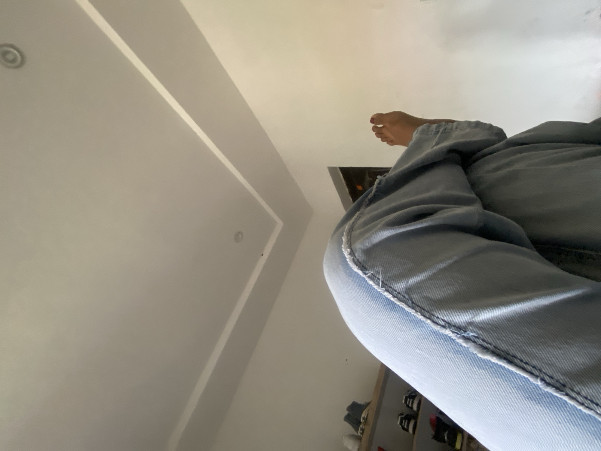 The image is taken from a low angle, looking up at a person wearing light-wash denim jeans and a visible foot with red nail polish on the toes. The person is standing indoors, as indicated by the white ceiling with recessed lighting. In the background, a partially visible shoe rack with various pairs of shoes can be seen. There is no indication of the location being in Abuja, Nigeria.