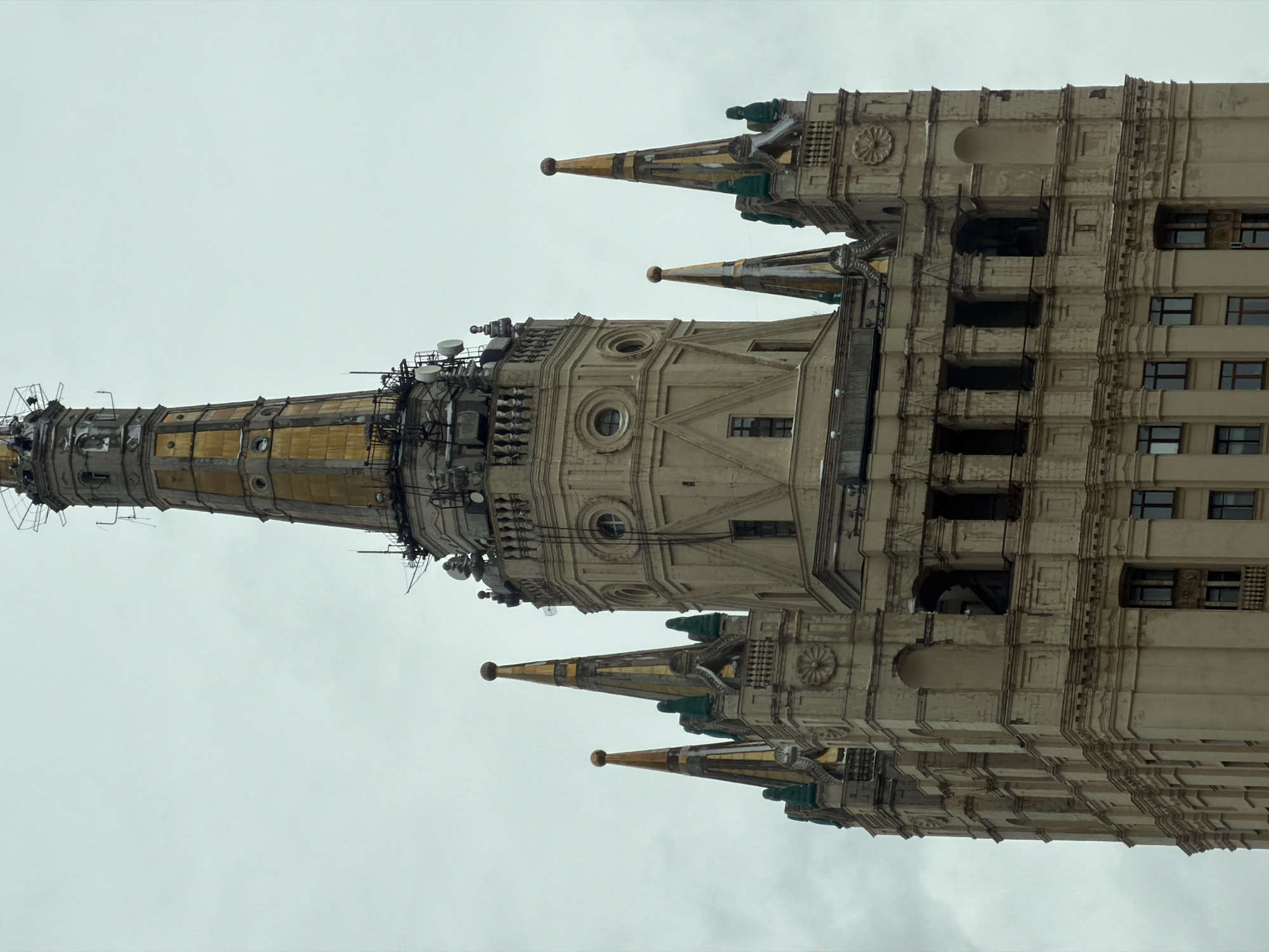 An exterior, vertical shot captures a grand, ornate high-rise building against an overcast, light gray sky. The building, characteristic of Stalinist Empire style architecture, is seen from a low angle, emphasizing its height and intricate details.
The central feature is a towering spire, which appears to be gilded or made of a golden-colored material, featuring multiple sections, railings, and platforms. Numerous modern communication antennas, satellite dishes, and other electronic equipment are affixed to this spire and the upper sections of the building, contrasting with its historical design.
Below the main spire, the building expands into a multi-story structure with a light, possibly off-white or beige, stone facade. This main body is adorned with classical architectural elements such as decorative pilasters, cornices, arched windows, circular windows, and intricate moldings including rosette patterns. Several smaller, decorative spires or turrets, also featuring gold and possibly green accents, protrude from the building's upper levels, adding to its palatial appearance.
The building features numerous rectangular windows, some appearing dark, suggesting reflections or interior shadows. Despite its grandeur, areas of the facade show signs of weathering, with some discoloration and apparent wear on the stone and gilded elements. No people or specific activities are visible, presenting a static architectural portrait. The overcast weather suggests diffused lighting, and the architectural style strongly indicates a location in Moscow, Russia. No visible text is present.