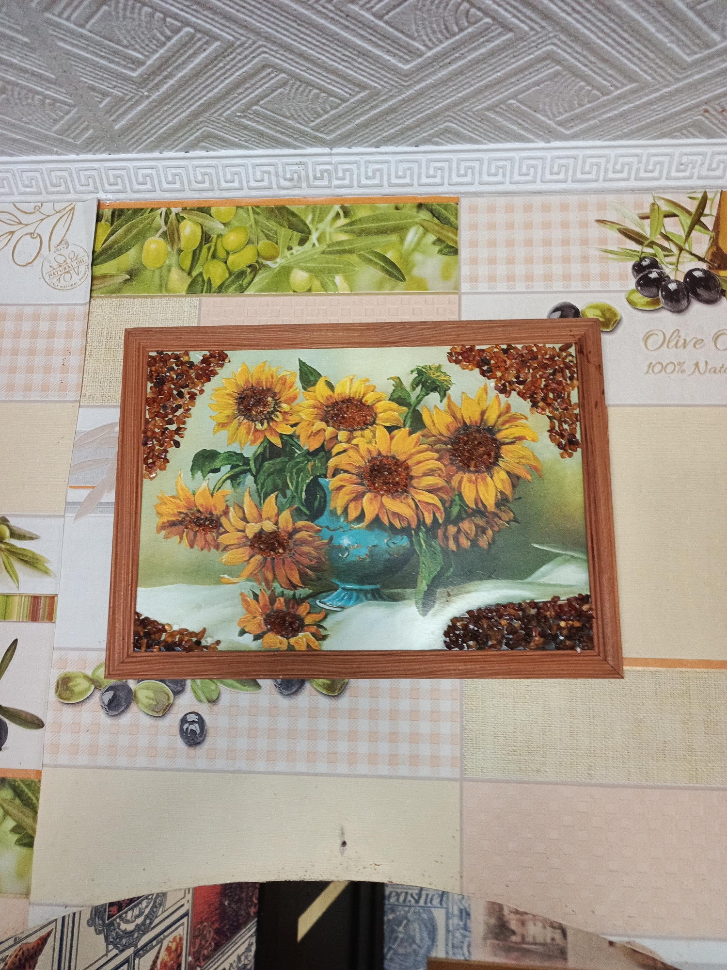 The image shows a framed painting of sunflowers in a blue vase, adorned with what appears to be amber pieces around the frame. The painting is hanging on a wall covered with a patchwork of tiles, some with olive motifs. The ceiling above has a patterned texture and a decorative molding. The photo was taken indoors, with details of a cabinet or counter visible at the bottom. The location is indicated to be in Chaikovsky, Russia, but the image itself does not provide any specific local landmarks.