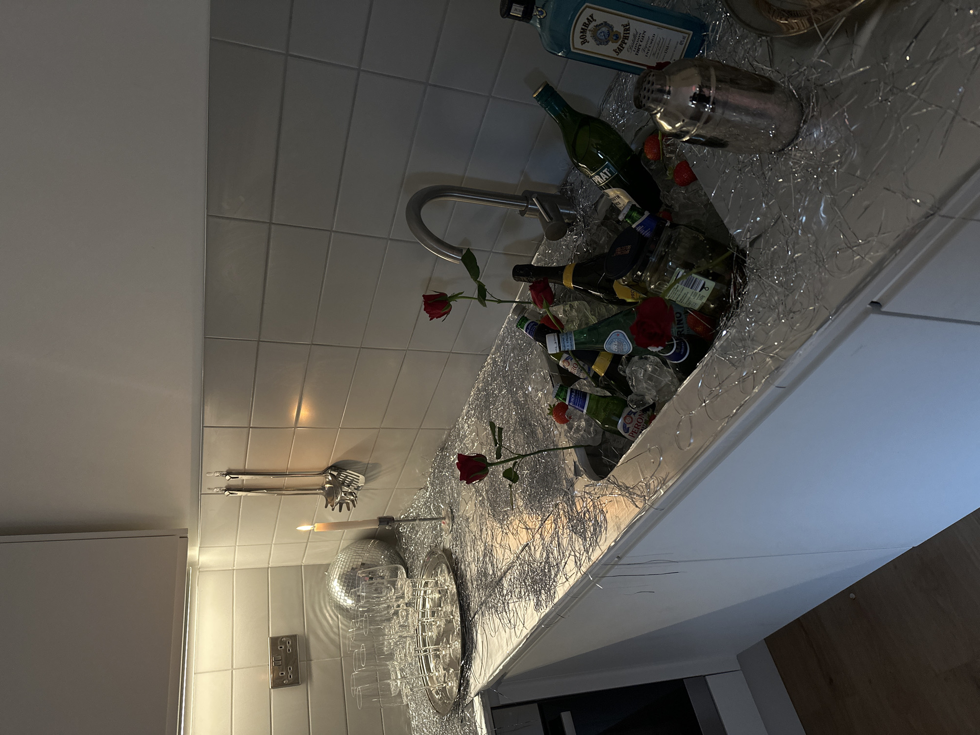 The image shows a kitchen sink filled with bottles of alcohol, ice, and roses, adorned with silver tinsel. A bottle of Bombay Sapphire gin is visible near a silver cocktail shaker. Other bottles include Peroni beer and wine. A silver faucet extends over the sink. Behind the sink, the wall is tiled. To the left of the sink area, kitchen utensils are mounted above a disco ball and a tray of glasses. A candle is lit nearby. The environment is domestic, likely a home kitchen decorated for a celebration. The location mentioned in the bottle label is London, United Kingdom.