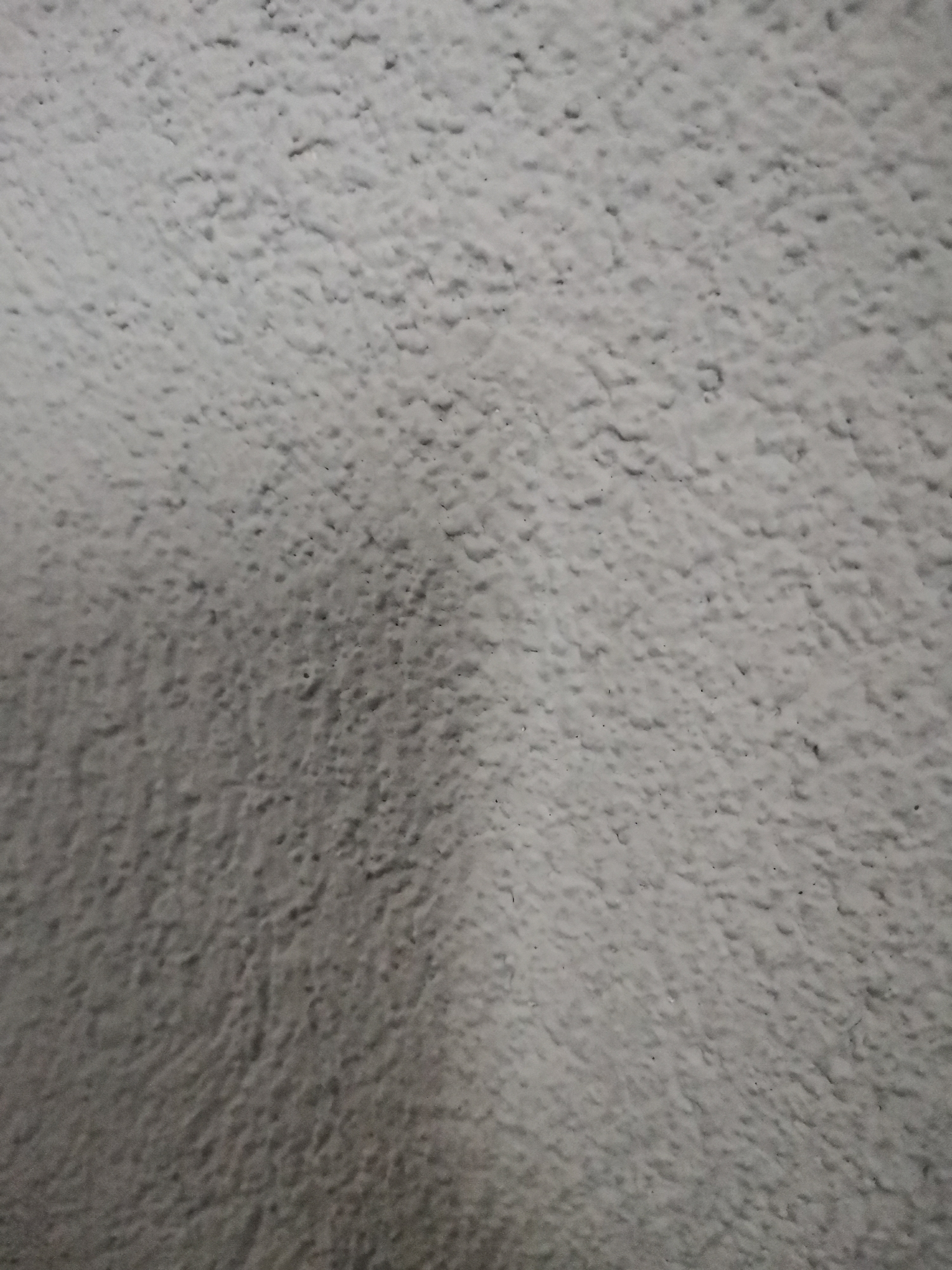 The image shows a close-up of a roughly textured surface, possibly a wall. The texture is uneven and appears to be painted or coated with a light grey material. The lighting is soft and directional, creating shadows that accentuate the texture. The scene is located in Pokrovnik, Bulgaria.
