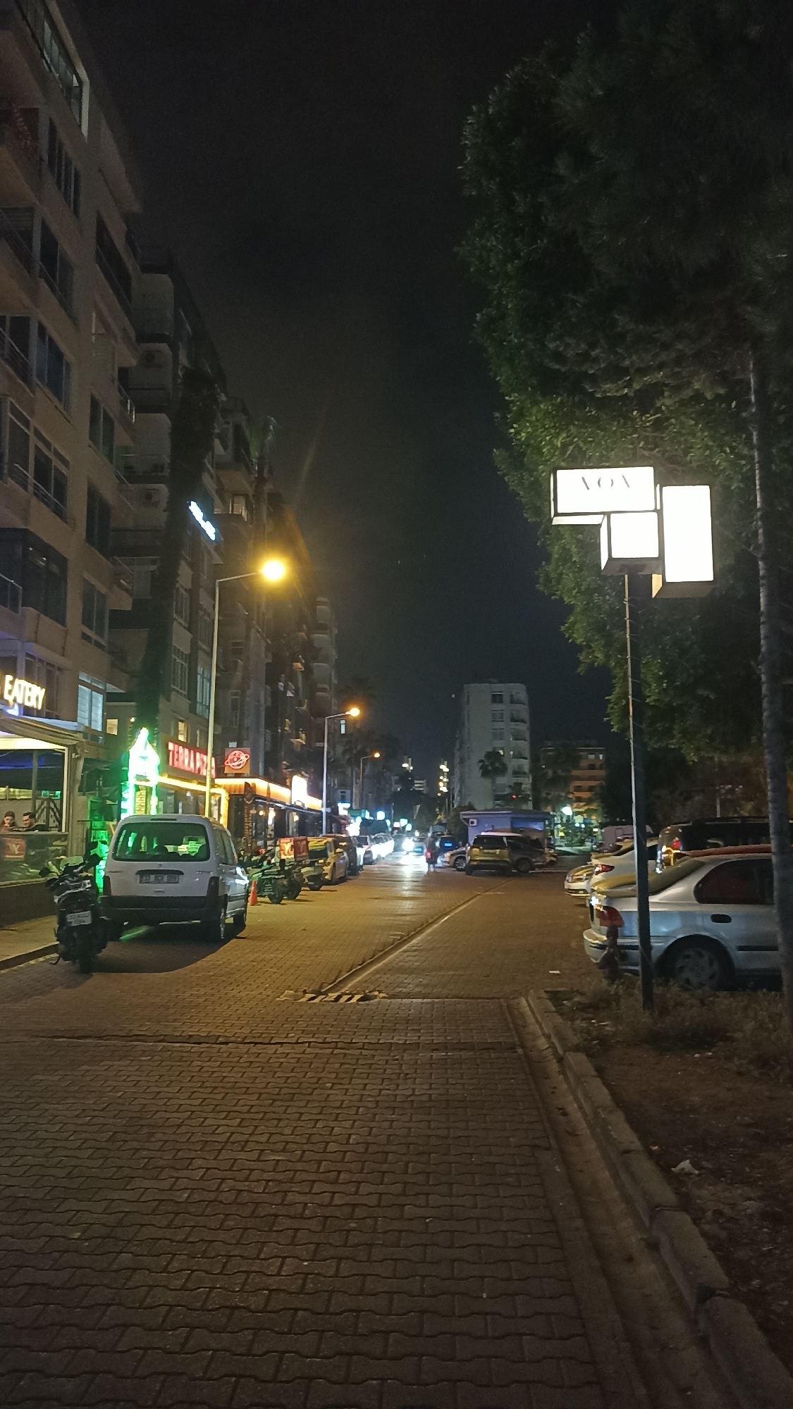 This nighttime street scene in Yenişehir, Turkey, depicts a paved road flanked by multi-story buildings and trees under a dark sky. Artificial lights from streetlamps, shop signs, and vehicle headlights illuminate the area. On the left, a row of buildings houses ground-floor businesses; a bright green neon sign reads "EATERY," and further down, another sign shows "TERRA PIZZA." Several cars are parked along the street, including a white car with a visible Turkish license plate (33 DCH 960) and a motorbike in front of it.
The road surface in the foreground is patterned, resembling cobblestones or interlocking pavers, with a slight raised section. On the right side, a substantial tree stands alongside a sidewalk and a patch of ground. A notable street sign is mounted on a pole near the tree, featuring multiple rectangular light boxes, with the word "VOA" clearly legible on the top box. In the distance, more buildings and vehicle lights are visible, receding into the night. A few people can be seen inside the "EATERY." The scene conveys a calm, urban atmosphere during the evening or night.