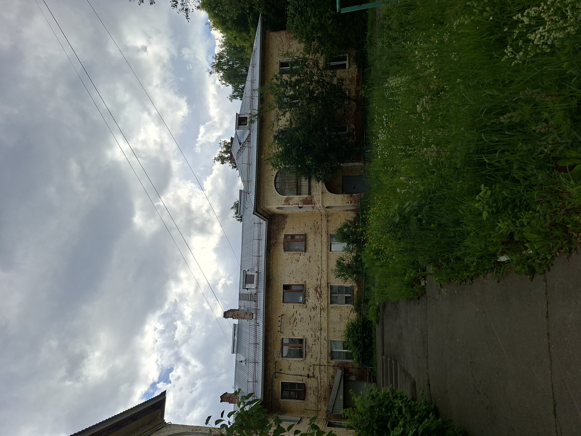 The image depicts the exterior of a weathered, three-story apartment building with pale yellow brick walls, likely in Izhevsk, Russia. The building has a metal roof and numerous windows, some of which appear to be boarded up or in disrepair. The architecture suggests it could be a Soviet-era residential structure. In front of the building, a concrete pathway leads to an entrance. The area is overgrown with tall grass and wildflowers, indicating a lack of recent landscaping. Trees and foliage surround the building and extend into the background, suggesting a semi-urban or park-like setting. The sky is overcast with dramatic clouds, implying inclement weather. There are no people visible in the frame. Power lines are visible crossing the sky.