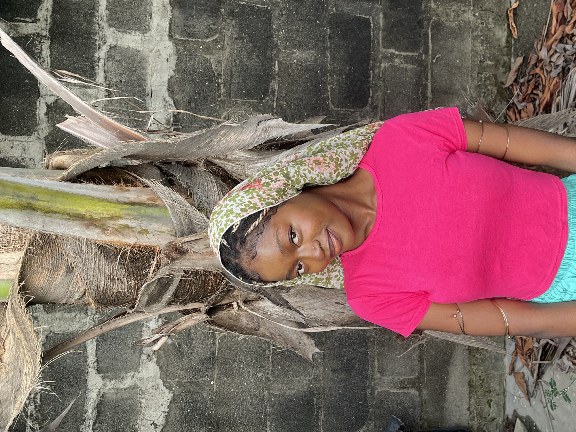 An eye-level shot captures a young woman posing next to a textured tree trunk and a concrete block wall, likely outdoors in Ado, Nigeria. The image is rotated, presenting the woman as if lying down, but she appears to be looking directly at the camera with a slight, friendly smile.
The woman has dark skin and wears a vibrant, short-sleeved pink t-shirt and light blue shorts. Her hair is braided, and a patterned headscarf featuring green, white, and pink floral or leaf motifs covers part of her head. She has a small stud piercing on her nose and multiple golden arm cuffs or bracelets on her right arm.
She is positioned close to the base of a large tree, possibly a palm, characterized by its rough, fibrous bark and several dried, fallen fronds or leaves. In the background, a wall constructed from grey concrete blocks is visible, showing a somewhat weathered texture. The lighting suggests it is daytime. No text is visible in the image.