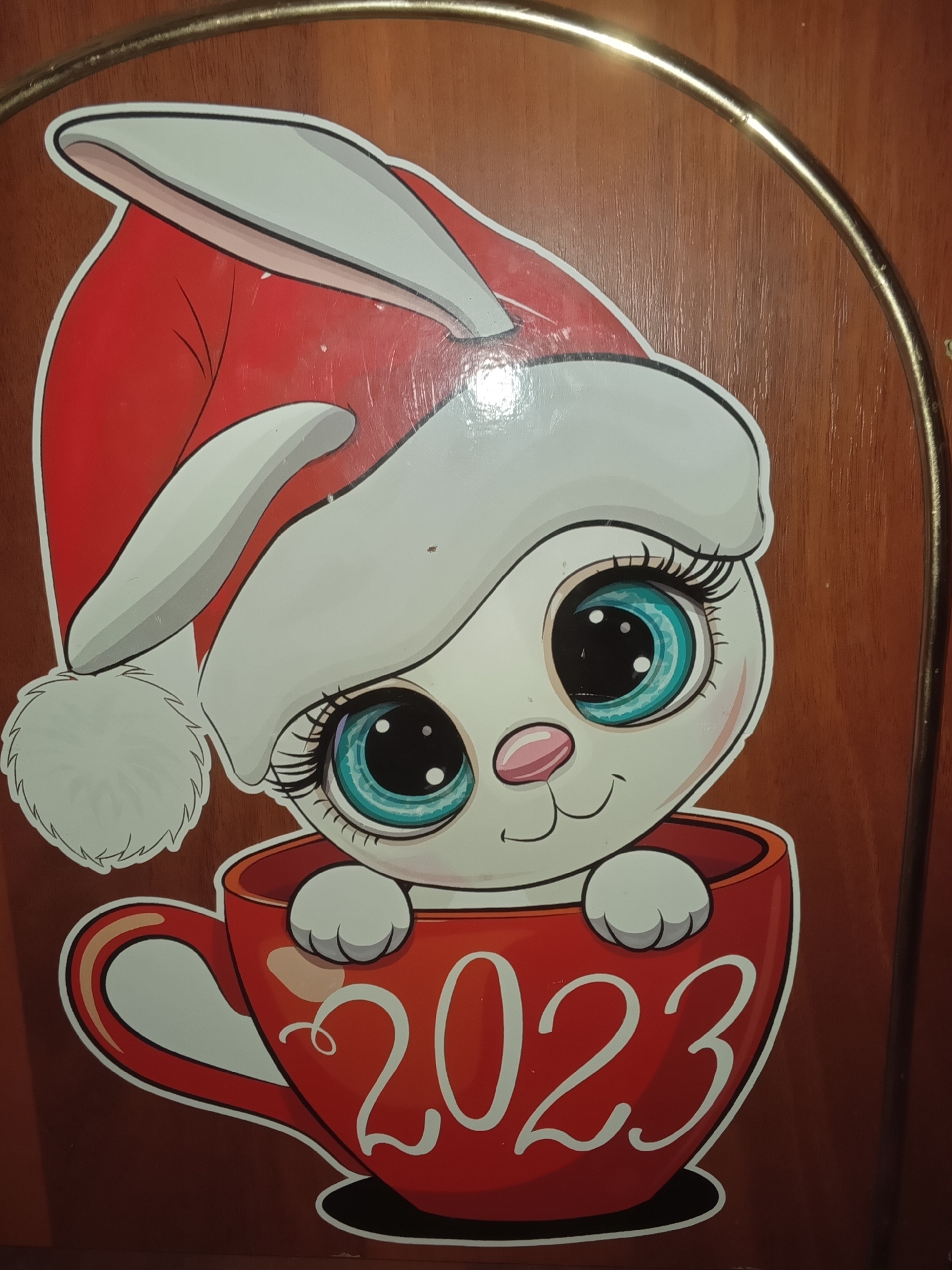 A photograph shows a sticker of a white rabbit wearing a Santa hat, peeking out of a red cup with the number "2023" on it. The sticker is affixed to a dark wooden surface, possibly a door or piece of furniture. A gold-colored metal hoop is visible in the upper right and top left corners of the image, suggesting the sticker is attached to a piece of furniture with a metal frame.
The photograph was taken at an unknown location in the city of Puchezh, Russia.