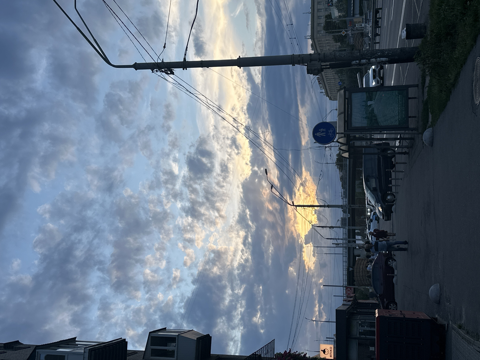 The image depicts a street scene in Kyiv, Ukraine, at dusk. The sky is filled with dramatic clouds, with a sliver of sunset visible through them. On the left, a building with balconies juts into the frame. A sidewalk runs along the right side of the street, with a bus stop shelter and a light pole. Several cars are visible on the street, and a few pedestrians are walking on the sidewalk. The overall mood is serene, with the soft light of dusk casting long shadows. The architectural style of the buildings and the presence of Cyrillic script on signage suggest a Ukrainian setting.