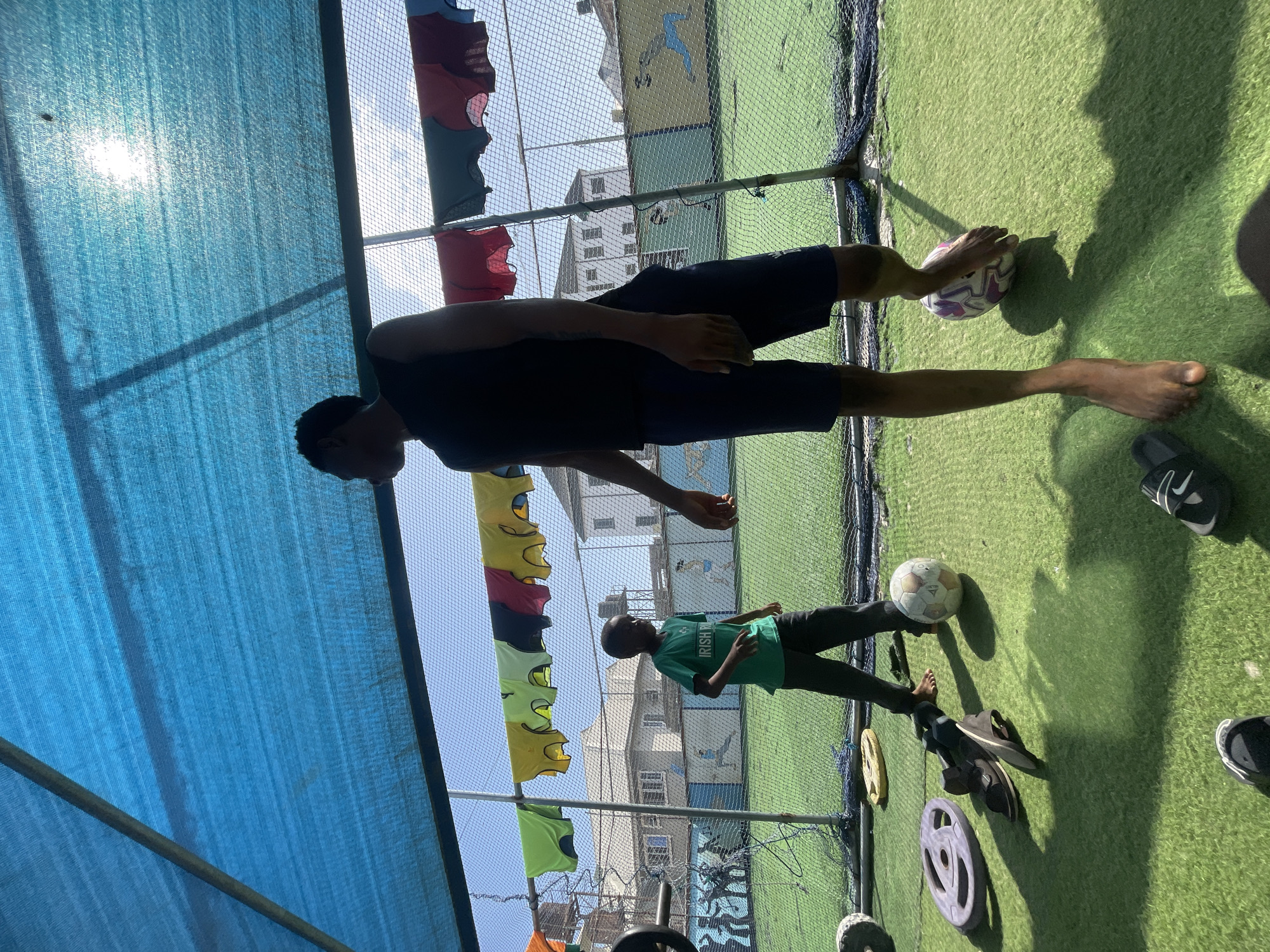 The photograph captures an outdoor scene at what appears to be a sports facility in Iranla, Nigeria, likely an artificial turf soccer pitch, under bright daylight. The image is oriented vertically but depicts the subjects horizontally on the ground.
In the foreground, a young adult male with a dark complexion stands on the left, wearing a dark t-shirt and dark shorts. He is barefoot, with one foot near a white and purple soccer ball. His face is largely in shadow. On the right, a child, also with a dark complexion, stands barefoot on the green artificial turf. The child wears a green t-shirt with the text "IRISH PUB" visible across the chest, and dark pants. Another white soccer ball is visible near the child's feet, along with sandals and disc weights scattered on the turf.
The background features a fence or netting separating the playing area from several multi-story buildings under a clear, bright sky. Colorful sports bibs or jerseys hang on a line across the fence. A blue mesh shade net covers the upper left portion of the scene, with the sun's glare visible through it, casting strong, distinct shadows of the subjects and structures onto the turf. The overall impression is a casual outdoor activity, possibly soccer practice or playtime, on a sunny day.