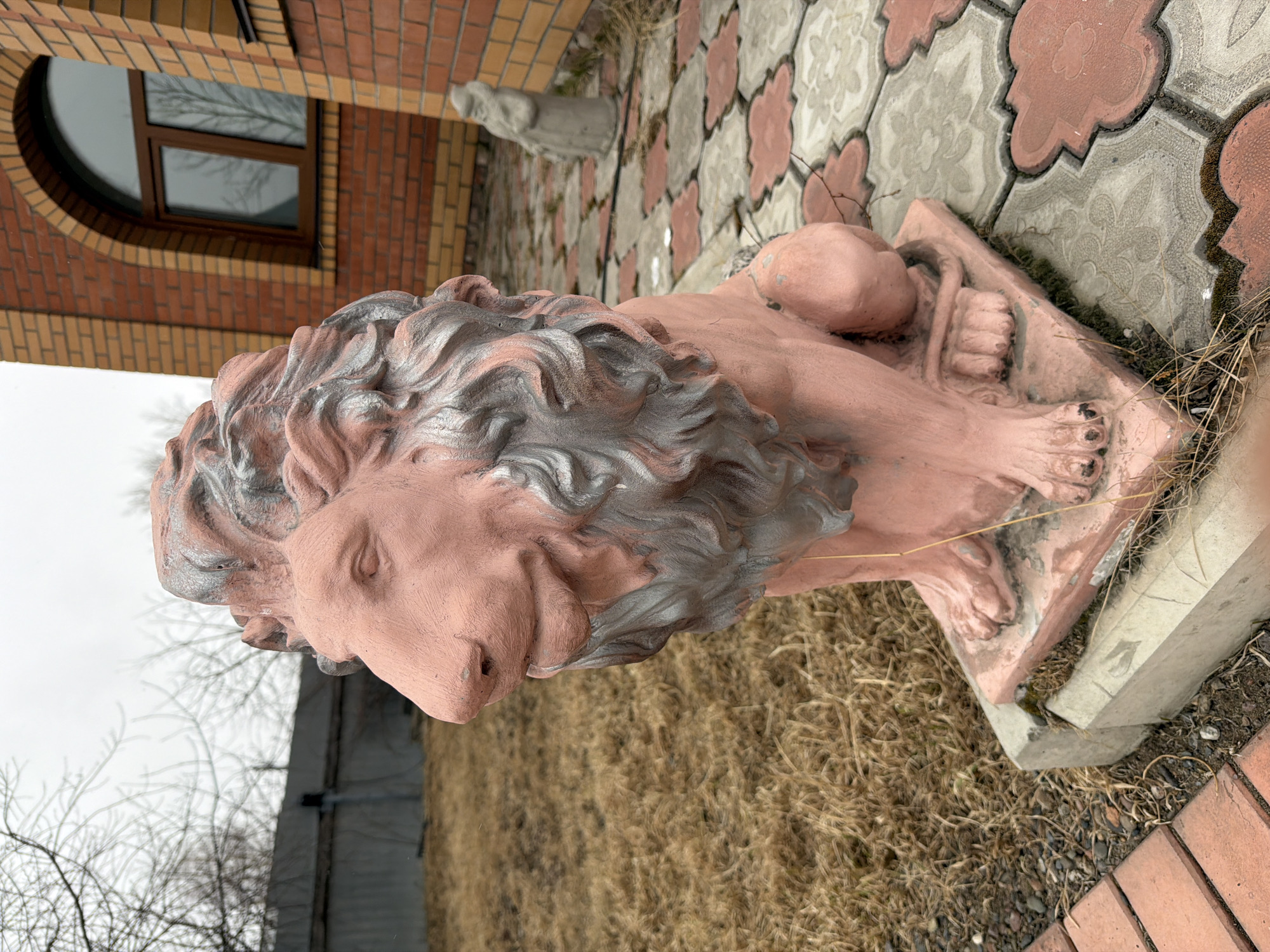A color photograph, taken outdoors during daylight hours, depicts a reclining lion sculpture. The sculpture is painted a reddish-pink hue, with a gray mane. It is positioned on a light gray, square base. The lion is located near a brick building with a round window and an attached gargoyle. The background features a pattern of red and gray cobblestones. The scene appears to be located in Krasnoyarsk, Russia. Brown dry grass and bare trees are also visible in the image.