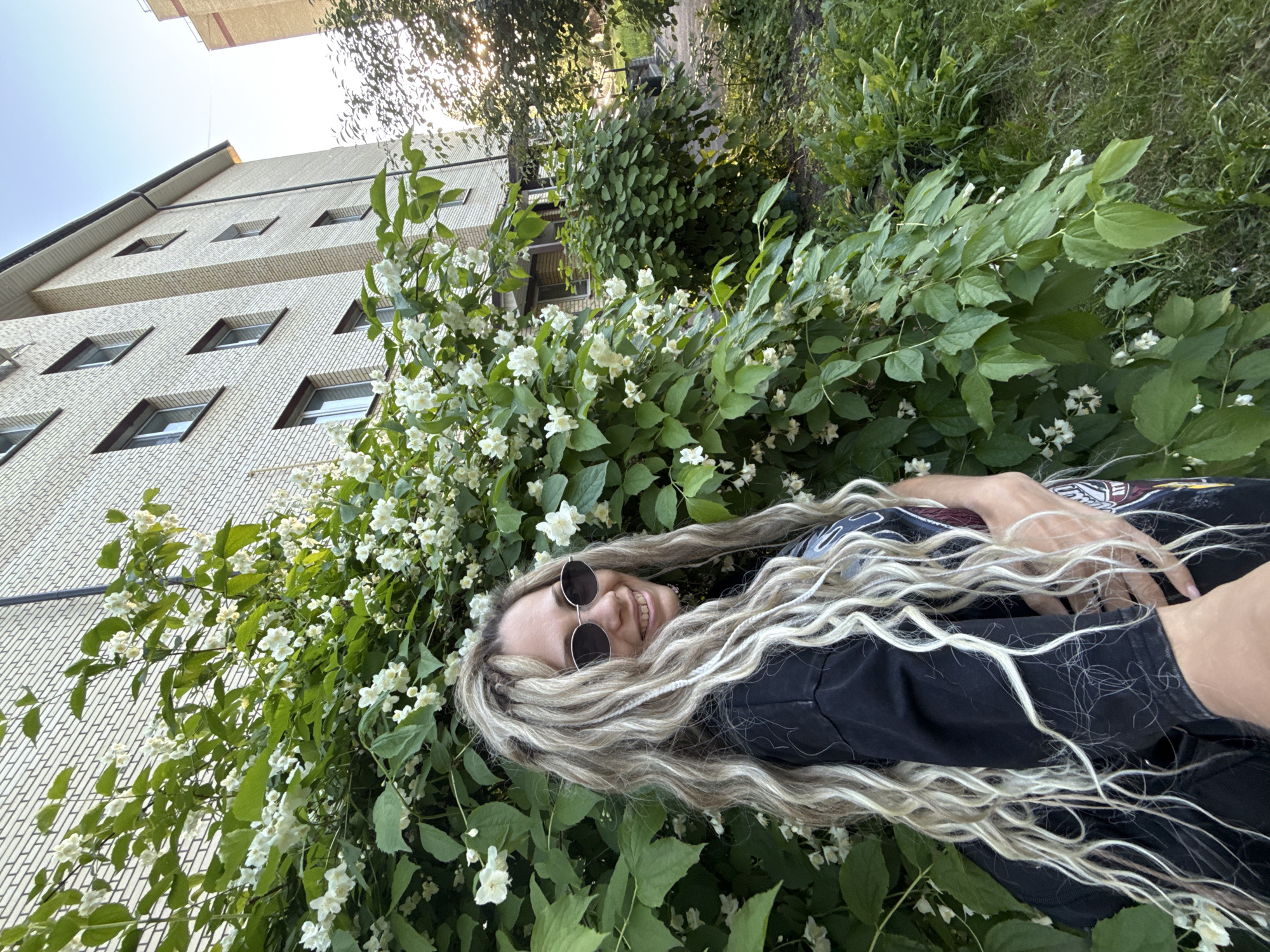 This image depicts an outdoor scene in the city of Rusanovo, Russia. A female subject is positioned in the left-center of the frame, facing slightly towards the right and looking directly at the camera with a smile, revealing her teeth. She has long, wavy hair characterized by a blend of blonde, light brown, and braided/dreadlock-like strands. She wears dark, oval-shaped sunglasses and a black, long-sleeved shirt with a partially visible graphic design on the chest area. Her right arm is extended forward, suggesting she is holding the camera or being photographed at close range, while her left hand rests on her waist.
Behind the subject and extending across the left and central midground is a large, dense bush covered in numerous small, white flowers and green foliage, likely a jasmine species. In the background, a multi-story residential building constructed of light-colored brick is visible, featuring multiple rectangular windows with dark frames. A portion of another, taller light-colored brick building is discernible in the far right background. Additional green bushes, grass, and trees are present in the midground and background, particularly to the right. The sky, visible in the upper right, is light blue, with a bright glare indicating the position of the sun, consistent with late afternoon or early evening lighting conditions.