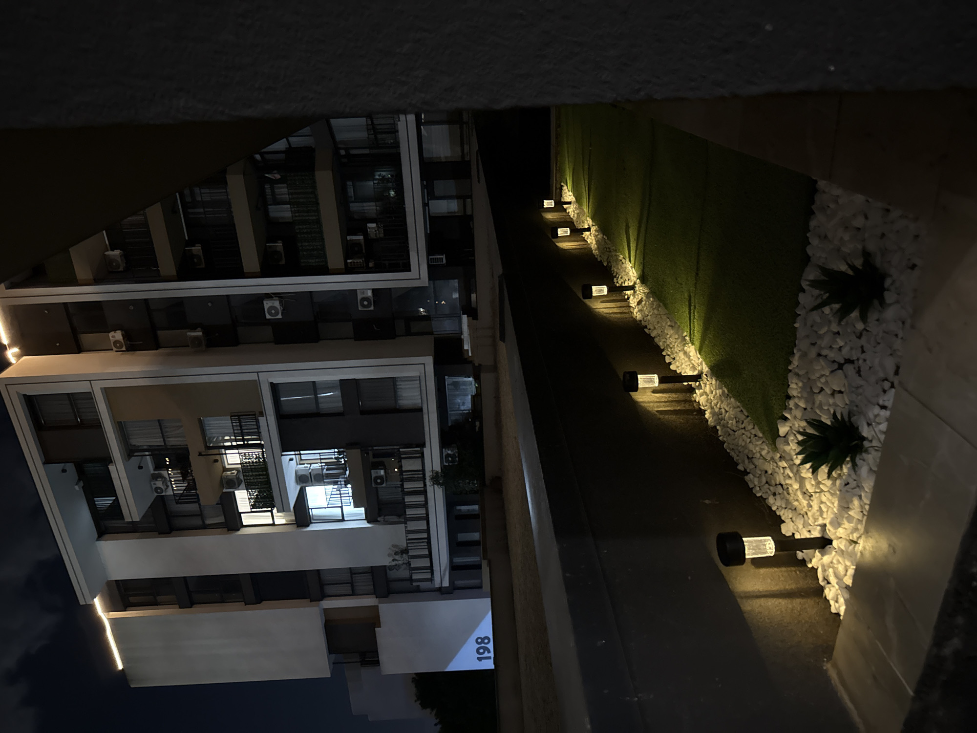 The image shows an exterior night scene. A multi-story building is visible, with the number "198" displayed on its side. The building features multiple air conditioning units and balconies. A path lined with small solar lights runs alongside the building. The path is adjacent to a strip of artificial turf bordered by white pebbles, and several succulent plants are interspersed among the pebbles. The location is inferred to be Burgas, Bulgaria.